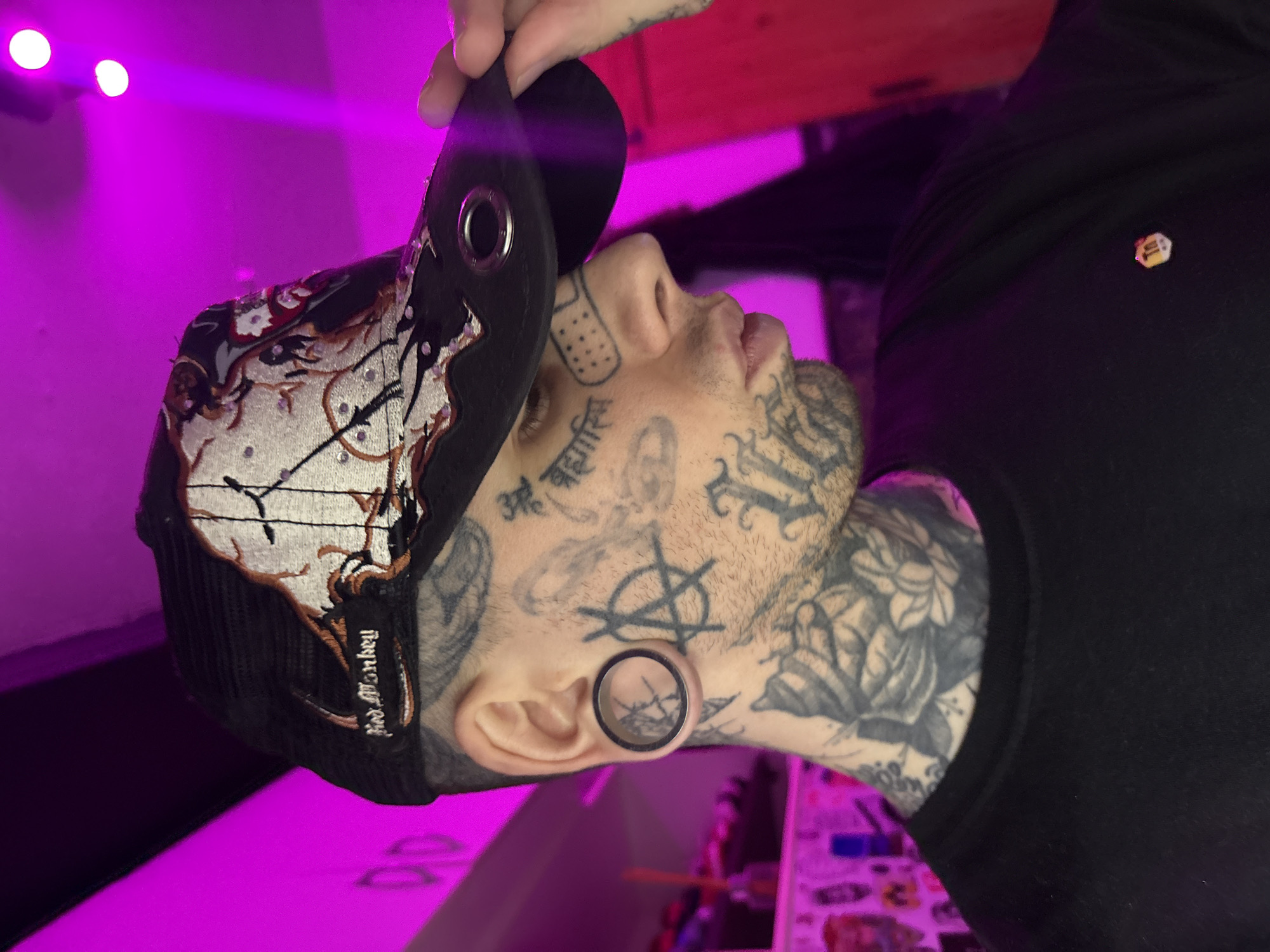 An individual, likely male, is depicted in a profile shot from the chest up, facing right and slightly downward. The person is extensively tattooed on the face and neck. Visible facial tattoos include an anarchy symbol on the right cheek, Devanagari script (appearing as "बहासि") below the right eye, and a "Band-Aid" design on the right cheekbone. The right ear contains a large, dark-rimmed ear tunnel/plug with a clear center. Floral and organic black ink tattoos cover the neck and extend onto the visible right shoulder.
The individual wears a black crew-neck t-shirt, adorned with a small, hexagonal yellow/orange pin featuring "Tn" in white on the right chest. A black trucker-style cap with a mesh back is worn. The cap's front panel is embroidered with intricate white, grey, orange, and black stylized designs, including abstract shapes and lines, accented by small metallic studs. A "Red Monkey" label in white script is on the cap's left side. The brim of the cap, which features two silver-colored eyelets, is being held by the individual's left hand, with fingers visible.
The scene is an indoor urban environment within the city of Buenos Aires, Argentina, characterized by pervasive magenta/purple ambient lighting. The background is out of focus, showing two distinct bright light sources in the upper right. Partially visible elements on the left suggest shelving with items, and a vertical reddish-orange surface is present on the far right. The setting appears to be a studio, tattoo parlor, or similar establishment.