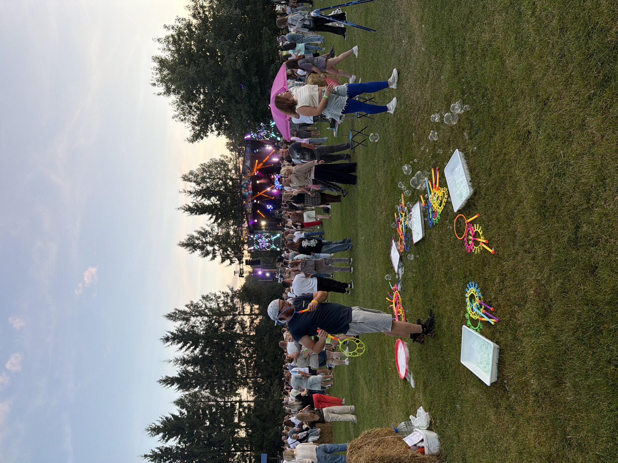 An outdoor festival is taking place on a grassy field with a line of trees in the background. A stage with lights and a band is visible in the distance. A crowd of people are milling about, some sitting and some standing. In the foreground, there are several tubs of bubble solution and bubble wands scattered on the grass. A man wearing a bandana and glasses is holding a bubble wand, appearing to be making bubbles. Other people are visible throughout the scene, some interacting with each other. The sky is a pale blue with a few clouds.