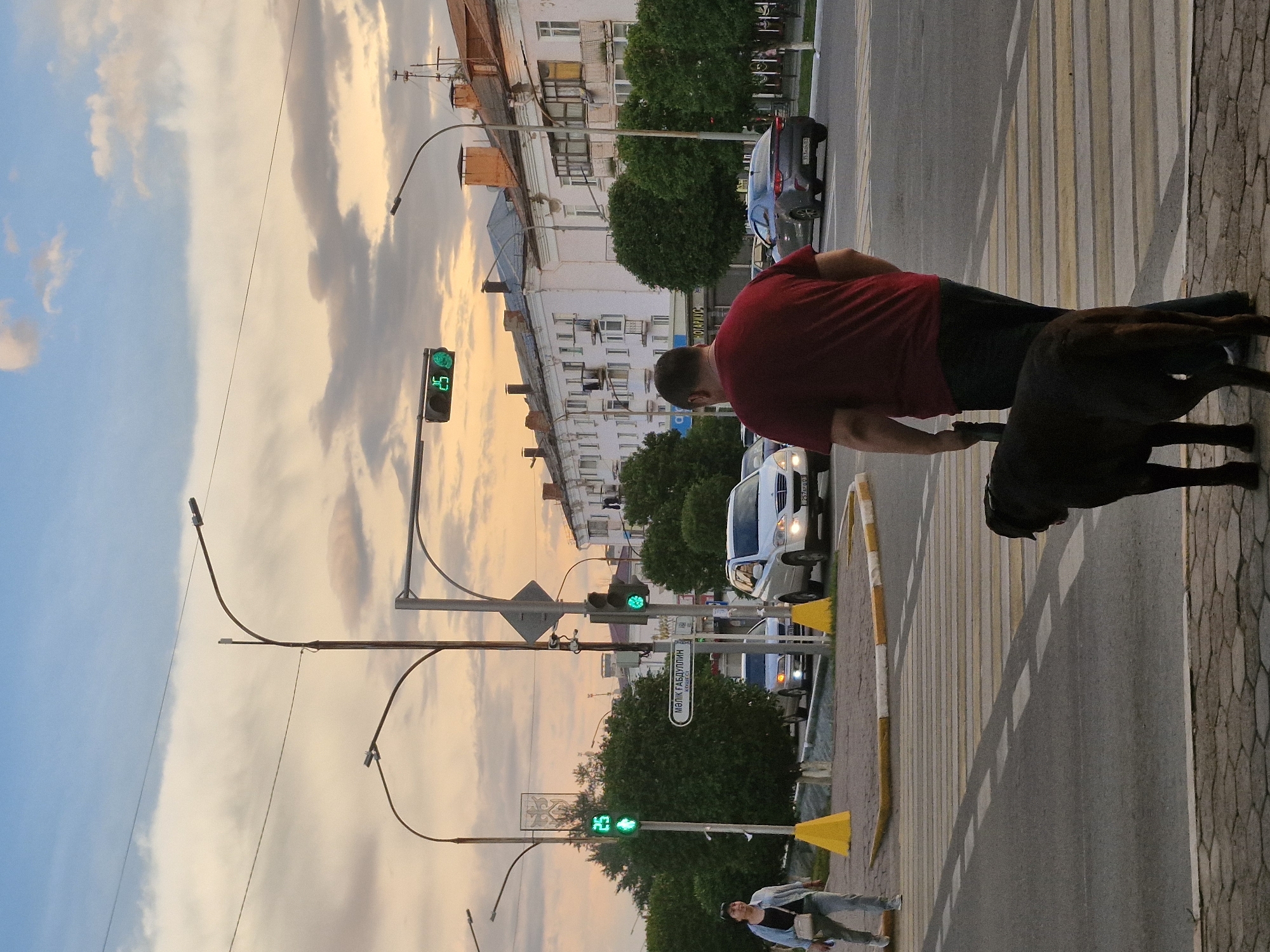 The image captures a street scene at dusk in Kokshetau, Kazakhstan, likely along Abylai Khan Avenue, a major thoroughfare. A man in a maroon t-shirt and dark pants is seen from behind, holding the leash of a dark-colored dog, possibly a Labrador, as they stand on a pedestrian crossing. Across the street, several cars are visible, including a white SUV and a dark sedan. Traffic lights are illuminated green, indicating that traffic can proceed. A woman in a light blue jacket and grey pants is walking on the sidewalk to the left of the frame, with a beige crossbody bag. Buildings line the right side of the street, featuring a mix of architectural styles. The sky is filled with dramatic clouds, tinged with the warm hues of sunset. The scene appears to be a typical urban environment during the transitional hours of the day.