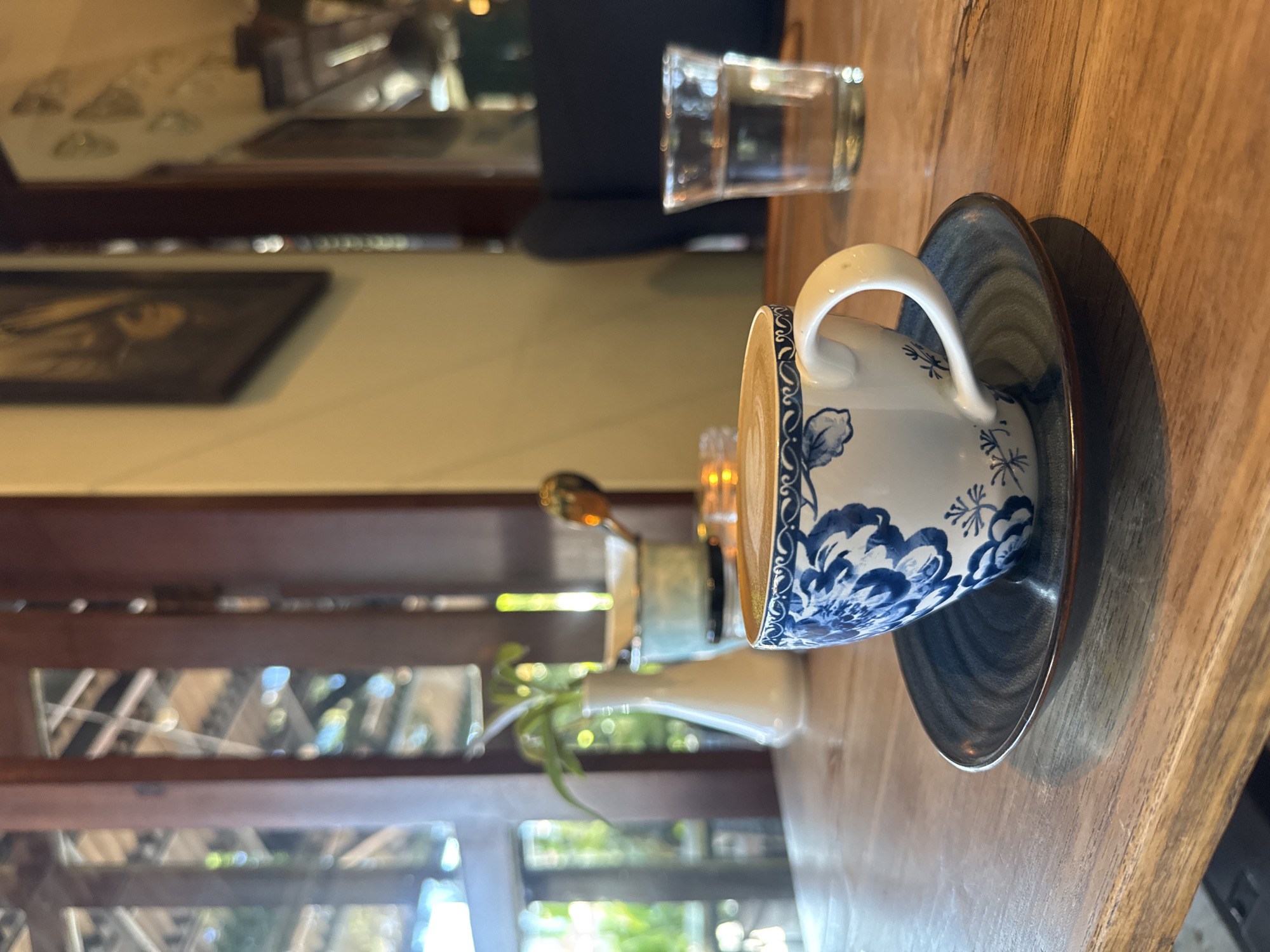 A cup of coffee with a blue and white floral design sits on a saucer on a wooden table. A glass of water is next to it. Through a window, greenery is visible. The location is North Kuta, Indonesia.