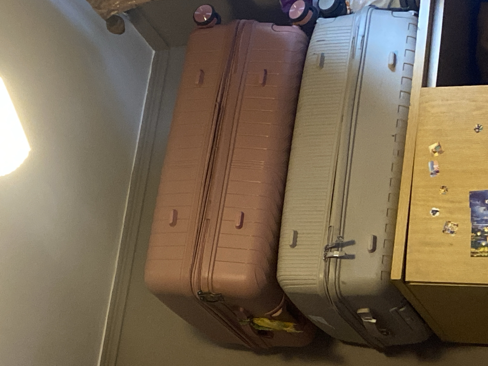 Two large suitcases are stacked on top of each other on top of a wooden cabinet. The bottom suitcase is a light grey color with ribbed detailing. The top suitcase is a muted pink color with similar ribbed detailing and a yellow tag hanging from one of its handles. The suitcases appear to be in a domestic setting, possibly a bedroom or storage area, with a wall and ceiling visible in the background. The scene is lit by an overhead light source, which is not fully visible in the frame. There are no people or animals present in the image. The overall impression is one of storage or preparation for travel.