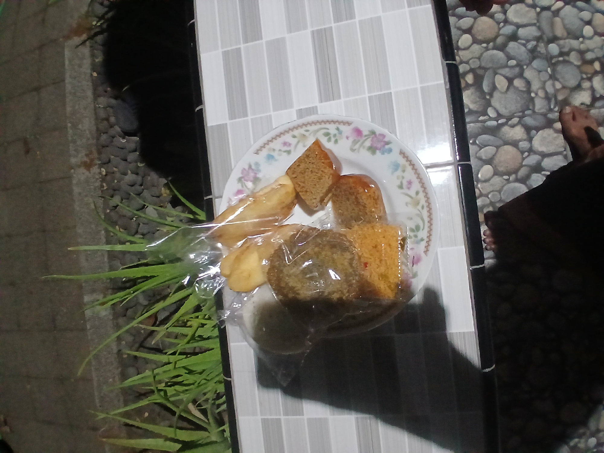 In Tenggarang, Indonesia, a plate of snacks sits on a tiled surface. The plate, adorned with floral patterns, contains several items, including what appears to be fried bread or pastries, some of which are cut into squares and covered in what looks like shredded coconut or breadcrumbs. A larger, round item, possibly a type of cake or bun, is also present and partially wrapped in plastic. To the left of the plate, a shadow suggests the presence of a person. In the background, a strip of green plants, possibly aloe vera, is visible against a dark, stony ground. The lighting suggests it is evening or night.
