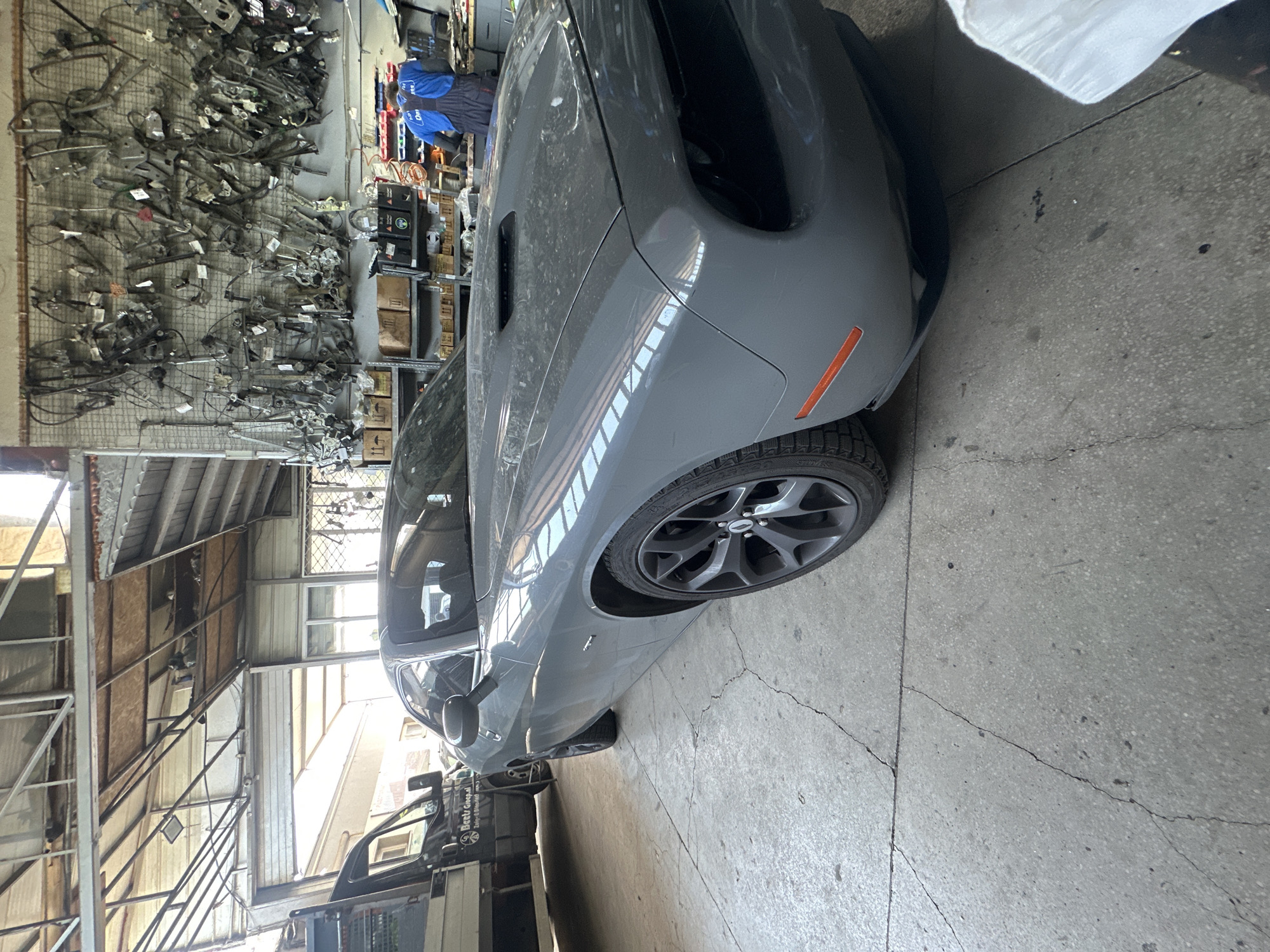In a dimly lit garage, a gray Dodge Challenger is parked on a concrete floor. The car is positioned at an angle, with its front end facing the right of the frame. The garage's ceiling is made of corrugated metal and exposed beams. On the right wall, a large collection of automotive parts, possibly window regulators, are meticulously organized and hung on a pegboard. Below the pegboard are shelves filled with cardboard boxes and various automotive supplies. A person wearing a blue work uniform with a white shirt underneath is standing near the shelves, facing away from the camera and towards the shelves, appearing to be working. In the background, a black truck is partially visible on the left side of the frame. The concrete floor is cracked and weathered, indicating a functional, working environment.