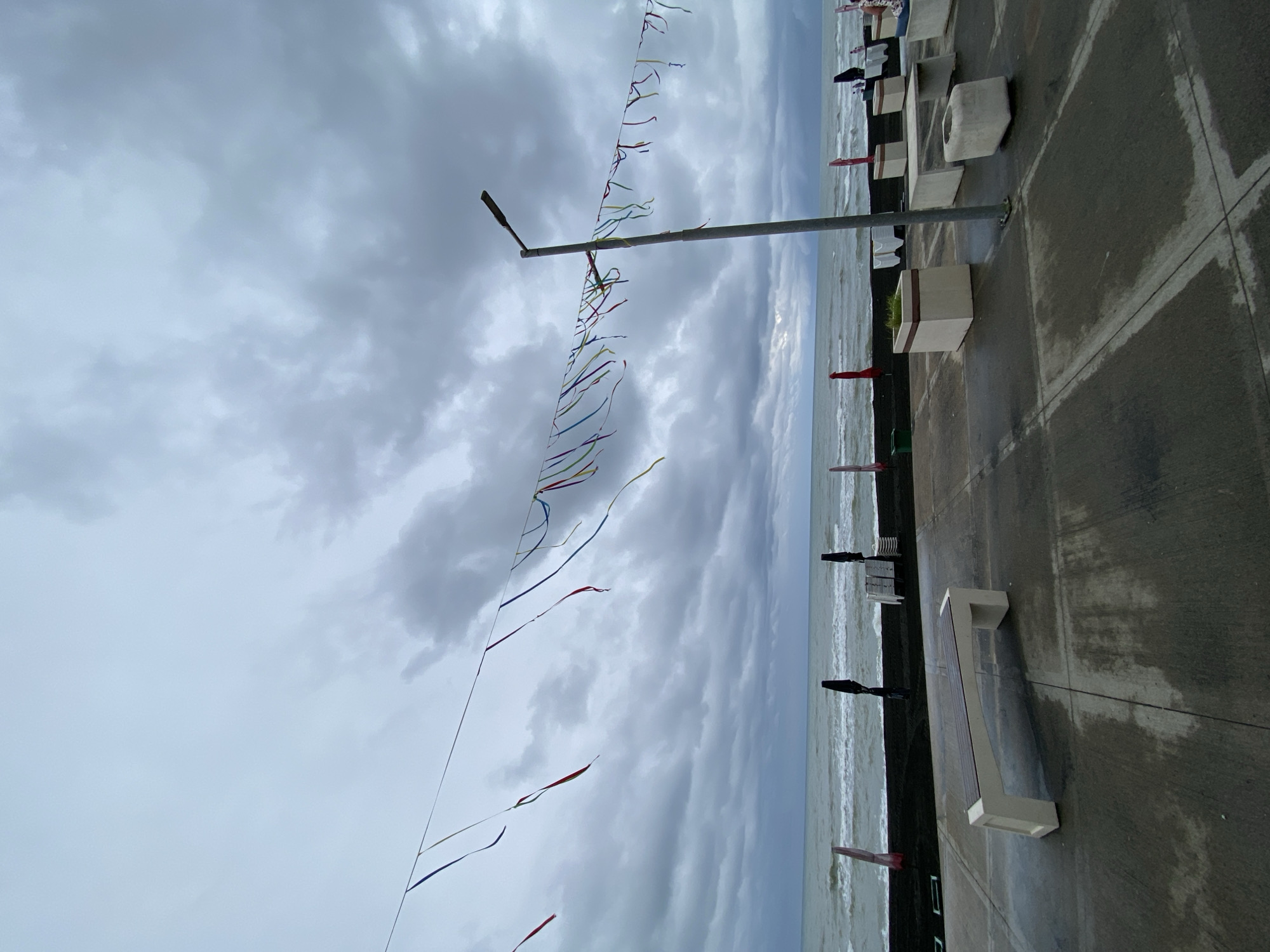 In Ureki, Georgia, on a cloudy day, a promenade faces the Black Sea. Colorful streamers are strung across the sky, attached to a lamppost. The promenade features concrete paving, wet from rain. A modern, cream-colored bench with a wooden seat is positioned on the left, and other concrete benches and planters are visible further down. Beyond the promenade, a dark beach borders the wavy, grey sea, with several black umbrellas and stacked white chairs suggesting a closed beach resort. The sky is overcast with heavy, dark clouds.