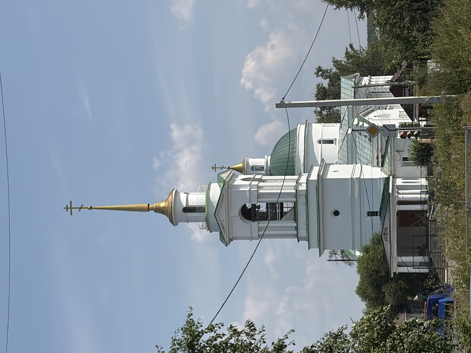 The image shows the Church of the Archangel Michael in Aramil, Russia. The church is a white building with a tall bell tower topped with a golden spire and cross. It has a green dome and is surrounded by trees. In the foreground, there are some power lines and a blue vehicle with an orange light on top. There are also some people visible near the entrance of the church. The sky is blue with some white clouds.