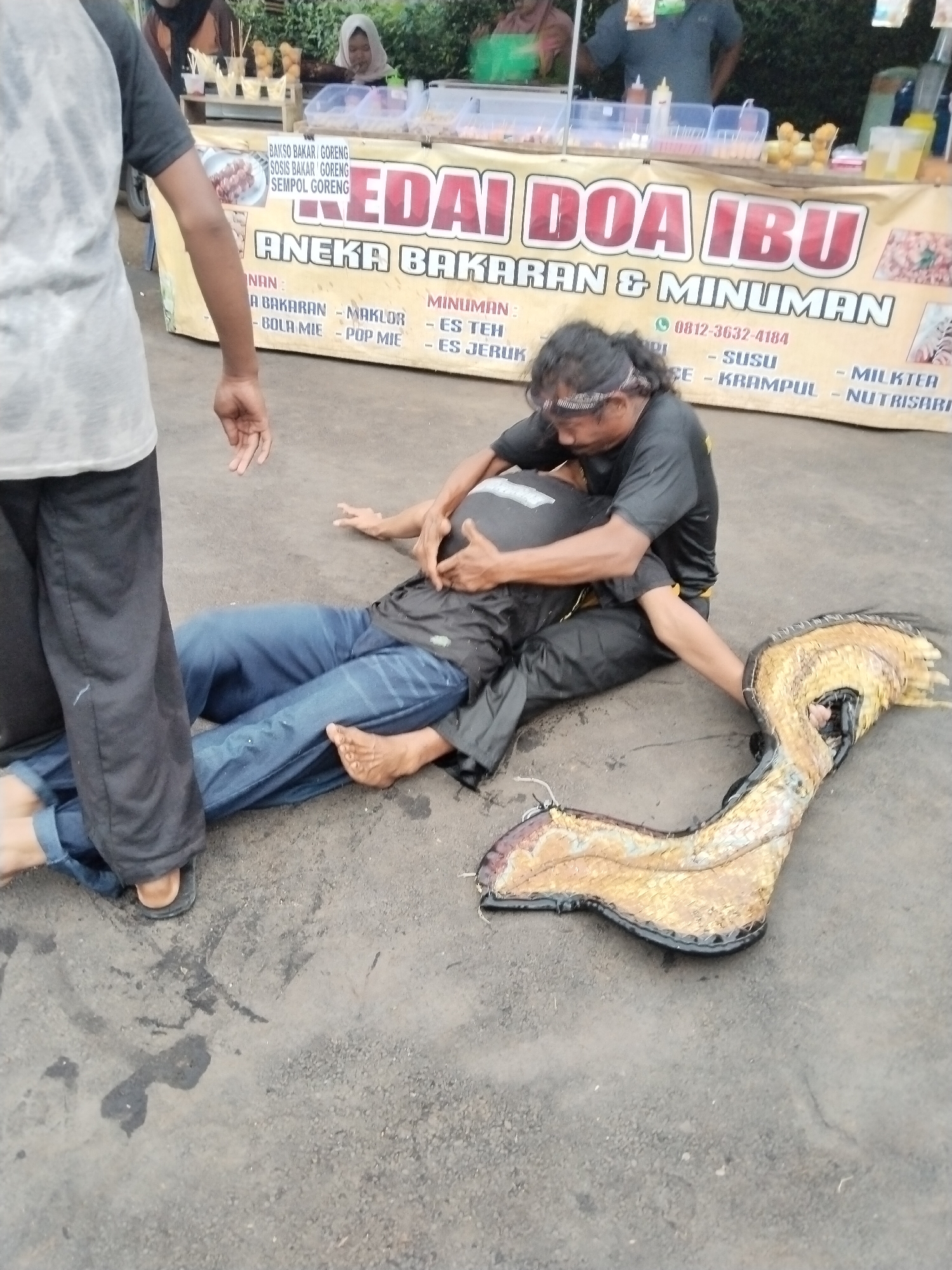 The image shows a street scene in Kuto, Indonesia. A man in a black shirt and pants is kneeling on the ground, seemingly providing some kind of treatment or assistance to another man who is lying on his back, wearing blue jeans and a black shirt. Another person is standing nearby, partially obscured on the left side of the frame. A large, decorative object resembling a sandal, with a gold-colored, scaly appearance, is lying on the ground beside the two men. In the background is a food stall with a sign reading "KEDAI DOA IBU ANEKA BAKARAN & MINUMAN," indicating it sells grilled foods and drinks. There are people behind the stall, possibly vendors or customers. The ground is paved and appears somewhat damp or stained.