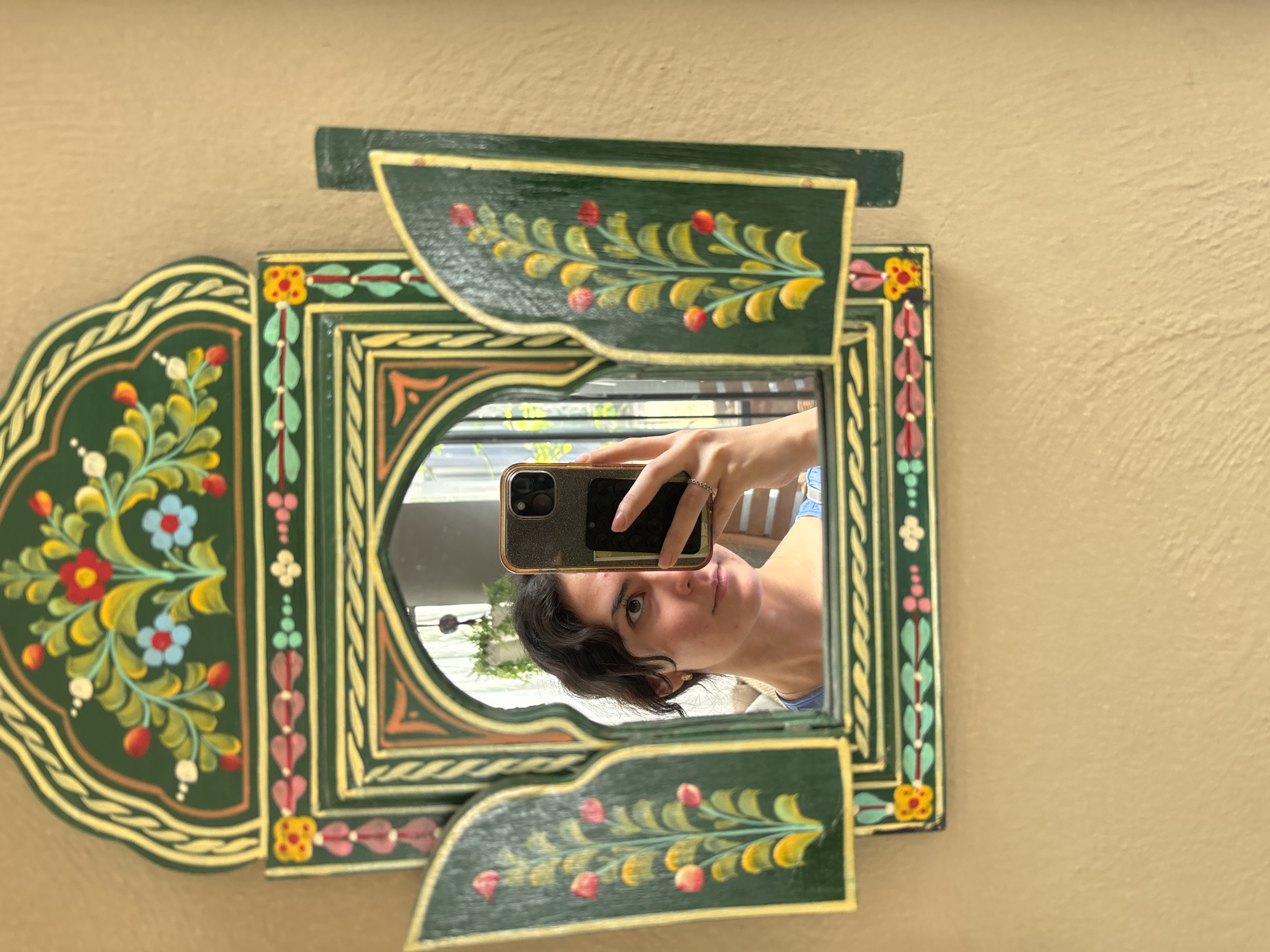 A woman is taking a selfie in a mirror. The mirror has a colorful, decorative frame, seemingly hand-painted with floral designs in red, yellow, green, and blue hues. The frame appears to be made of wood, and the background is a pale yellow or beige wall with a slightly textured surface. The woman is holding a cell phone in front of her face to capture the image, reflecting the room behind her, and she has short, dark hair and is looking at the phone. It appears the location may be in Puebla, Mexico.
