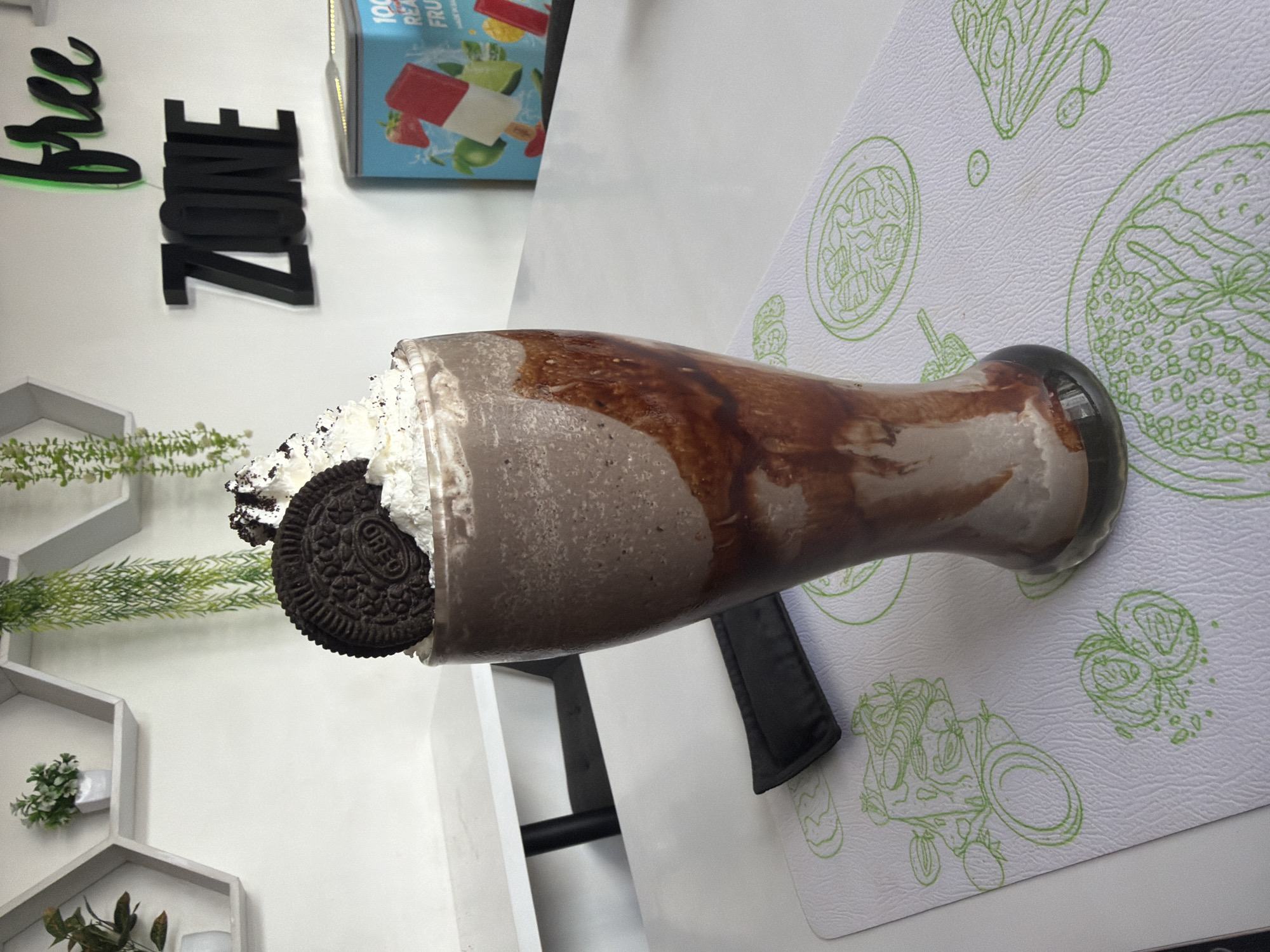 A tall glass of chocolate milkshake with whipped cream and an Oreo cookie is placed on a white table in a cafe in Mengwi, Indonesia. The milkshake has chocolate syrup drizzled down the sides of the glass. In the background, the words "FREE ZONE" are displayed on the wall in black and green lettering. There is also a cooler stocked with popsicles. The table has a white placemat with green line drawings of food items. The overall environment appears clean and bright, with white walls and shelves containing decorative plants.