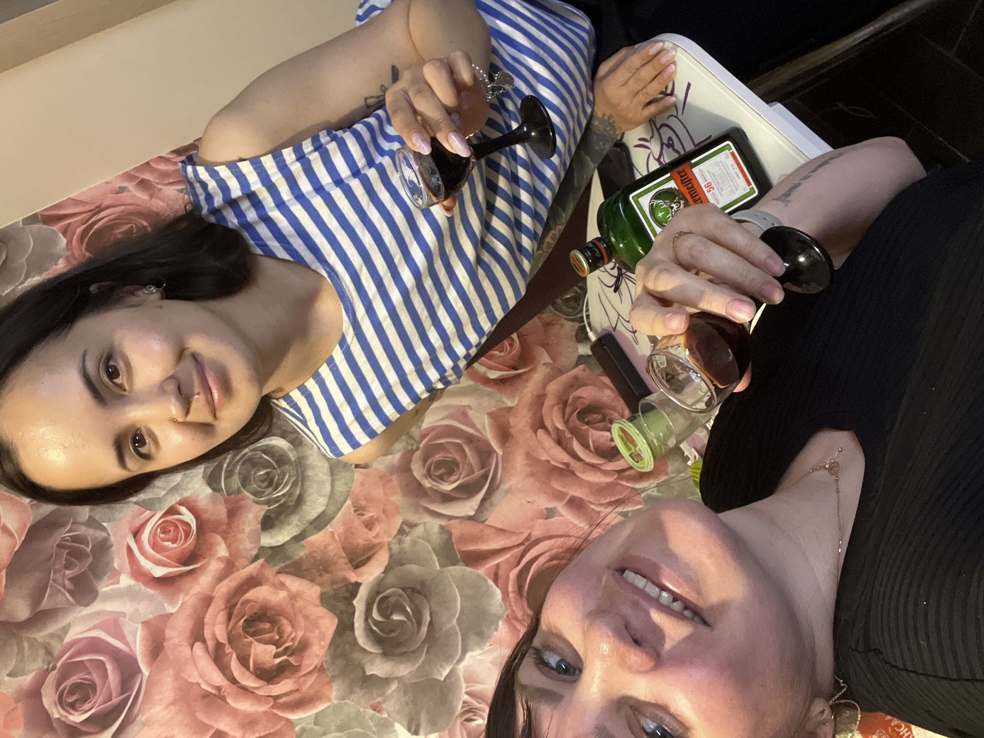 Two women pose with glasses of dark liquid and a bottle of Jägermeister. The woman on the left wears a blue and white striped shirt, while the woman on the right wears a black top. Both women are smiling. The background features a floral patterned surface, suggesting an indoor setting. A white tray is partially visible, holding the bottle and glasses. The location is Noviy, Russia.
