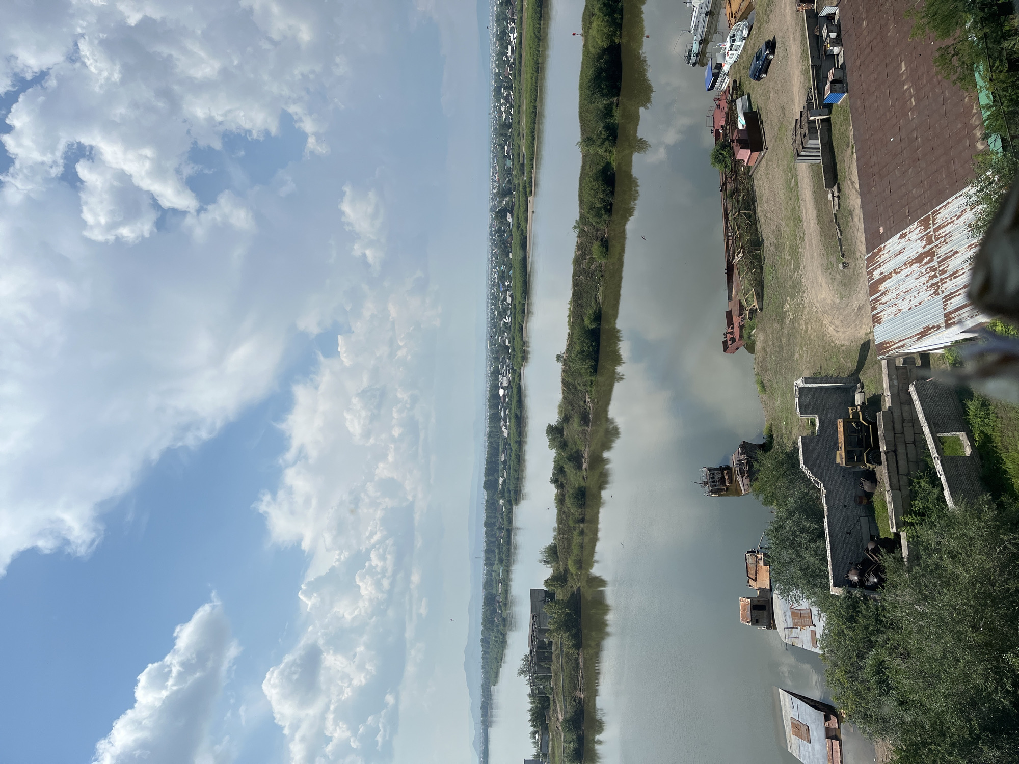 This is an aerial, wide-angle, eye-level shot of a river with a small industrial area on its bank, under a bright, cloudy sky. The river is wide and placid, reflecting the sky and surrounding greenery. On the right bank of the river, several derelict boats and barges are moored or partially submerged. There are also remnants of buildings, including a structure with a crumbling brick wall and a rusty, open-sided building with a corrugated metal roof. A light-colored utility vehicle is parked near a collection of industrial debris, including pipes and barrels. Further down the bank, a red barge with a crane-like structure is visible. Several smaller boats, including a white passenger boat and a dark blue vehicle, are also present. In the distance, across the river, is a densely populated town with numerous houses and buildings, nestled amongst green trees and backed by rolling hills. The sky is filled with fluffy white clouds against a clear blue backdrop. There are no people visible in the frame.