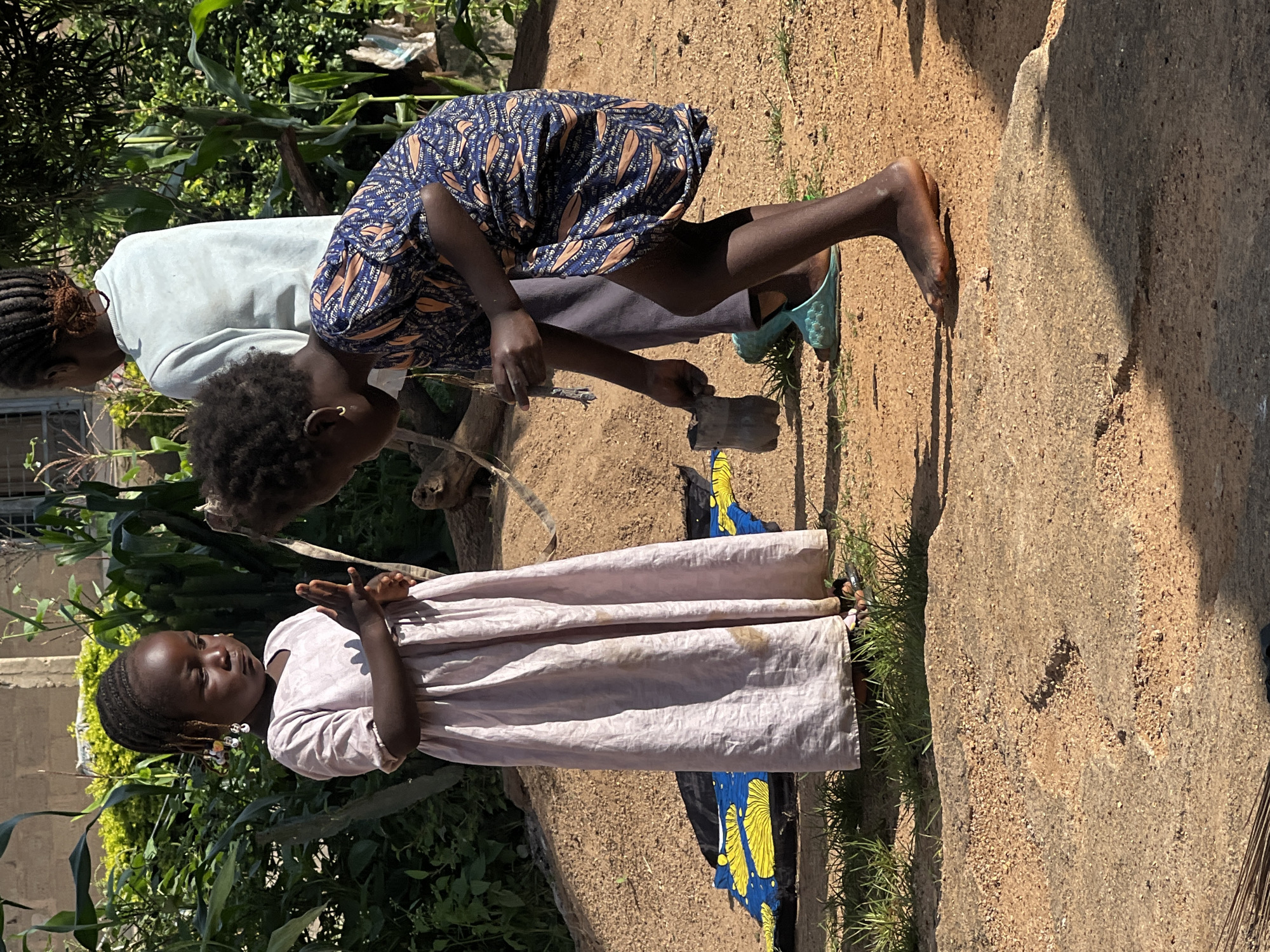 In Jos, Nigeria, three individuals are engaged in an activity outdoors, with a focus on two young girls in the foreground. The girl on the left, wearing a long, pale pink dress, stands facing forward with her hands clasped at chest height. She has braids adorned with colorful beads. To her right, a younger girl in a dark blue dress with an orange pattern is bent over, holding a dark object, possibly a piece of plastic or a small container, in her right hand. Her left hand is near the ground. Behind them, a third person, partially visible, wears a light blue long-sleeved shirt and gray pants. The setting appears to be a dusty, outdoor area with vegetation and a pile of sand in the background. The ground is a mix of dirt and gravel, with patches of grass. A colorful cloth with yellow and blue designs is spread on the ground to the left of the standing girl. The lighting suggests it is daytime, with shadows cast by the sun.