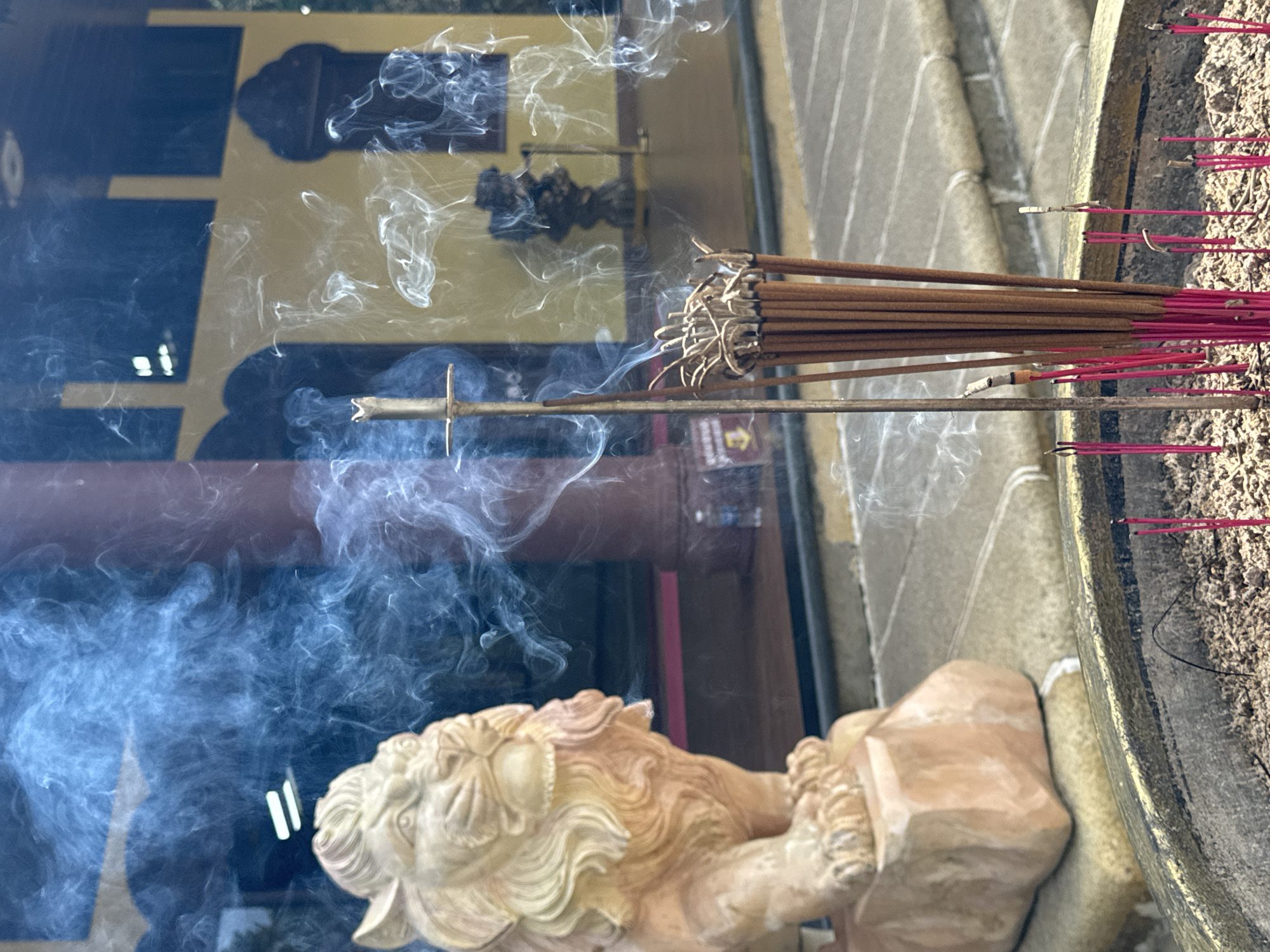 The image depicts a religious scene outside what appears to be a temple or pagoda in Da Lat, Vietnam. In the foreground, a large, weathered urn filled with ash and numerous incense sticks, some still burning and emitting smoke, is prominent. A brass holder for incense is positioned in the center of the urn, with a bundle of unlit incense sticks to its right.
To the left of the urn, a stone statue of a lion, carved in a traditional style, sits on a pedestal. The lion statue is light in color and weathered, suggesting it has been exposed to the elements.
In the background, a building with a bright yellow facade and dark arched windows is visible. The architecture suggests a Southeast Asian or East Asian influence. Smoke from the incense drifts upwards and across the scene, partially obscuring the background elements. The ground surrounding the urn and statue is paved with stone tiles. The overall atmosphere is one of reverence and tranquility. There are no people visible in the image.