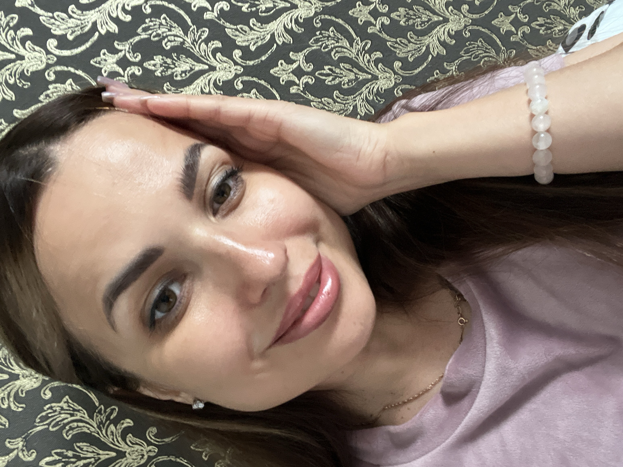 A medium shot, portrait style, shows a woman with dark brown hair and brown eyes, with her right hand touching her forehead. She is wearing a light pink sweatshirt and a delicate gold necklace with a small pendant. A pale pink beaded bracelet is visible on her left wrist. The background features a dark gray damask wallpaper with a repeating gold floral pattern. The lighting is soft and appears to be from the front, illuminating her face and highlighting her makeup, including lipstick and eye makeup.