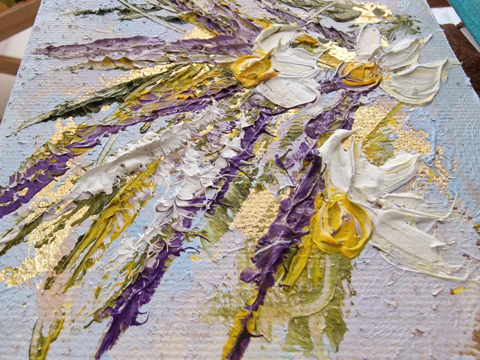 The image presents a close-up, angled view of a highly textured floral painting, located in Veliko Tarnovo, Bulgaria. The artwork features a vibrant, abstract bouquet created with a heavy impasto technique, where thick layers of paint produce a significant three-dimensional surface. Dominant colors include deep purple, bright yellow, and white, intermingled with olive and lime green hues for foliage. Numerous gold leaf or metallic flakes are embedded within the paint, scattering shimmering accents across the composition. The background of the painting is rendered in lighter tones of pale blue and lavender, also exhibiting visible brushstrokes and texture. Stylized petals and leaves are formed by distinct, raised strokes of paint. A brown and teal colored support, likely part of an easel, is faintly visible along the bottom right edge of the frame, indicating the painting is propped or displayed. No individuals are depicted.