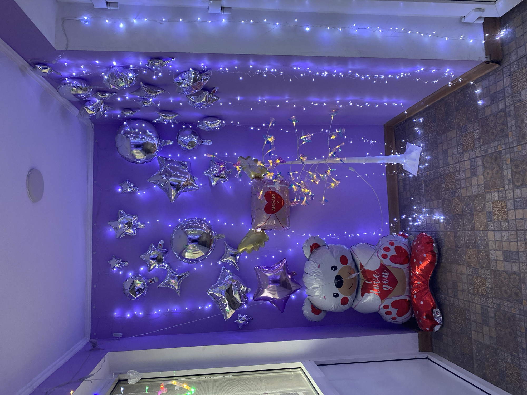 The image shows an indoor space decorated with balloons and fairy lights, possibly in Usolye-Sibirskoye, Russia. Several silver star and round balloons are attached to a purple wall and ceiling. A teddy bear-shaped balloon with the inscription "love you" and a red balloon in the shape of a heart. A decorative white branch with small lights is also present. Fairy lights are strung across the ceiling. The right side of the image shows a tiled wall and flooring, suggesting this area might be a hallway or a transitional space. A window is visible in the lower part of the image.