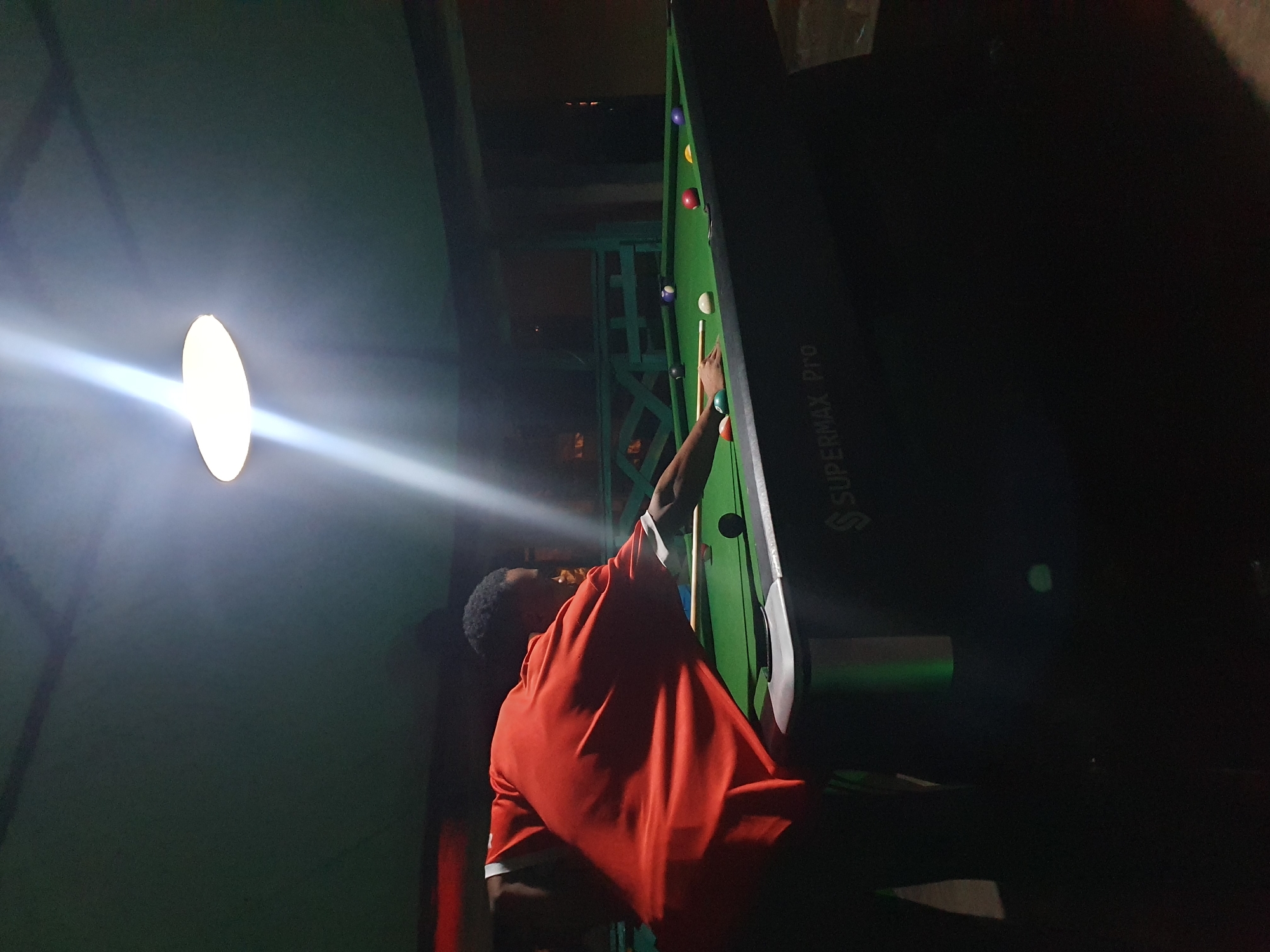In the dimly lit interior of a building, likely a recreation center or bar, a person is playing a game of billiards. The subject, a person wearing a bright orange t-shirt with white trim, is positioned at the end of a green pool table, cue in hand, ready to strike a ball. Several billiard balls, including a white cue ball, a red ball, and a purple ball, are visible on the table. The scene is illuminated by a bright, focused light source from the left, casting a strong beam across the table and the player. The background is mostly dark, with some structural elements visible, including what appears to be a metal railing or fence in the middle ground. A large, dark object, possibly a display screen or cabinet, stands to the right of the pool table, featuring white text that reads "SUPERMAX Pro". The overall atmosphere is one of focused activity in a somewhat atmospheric, possibly nocturnal setting. Given the context of Lokoja, Nigeria, it is likely an indoor, nighttime scene.