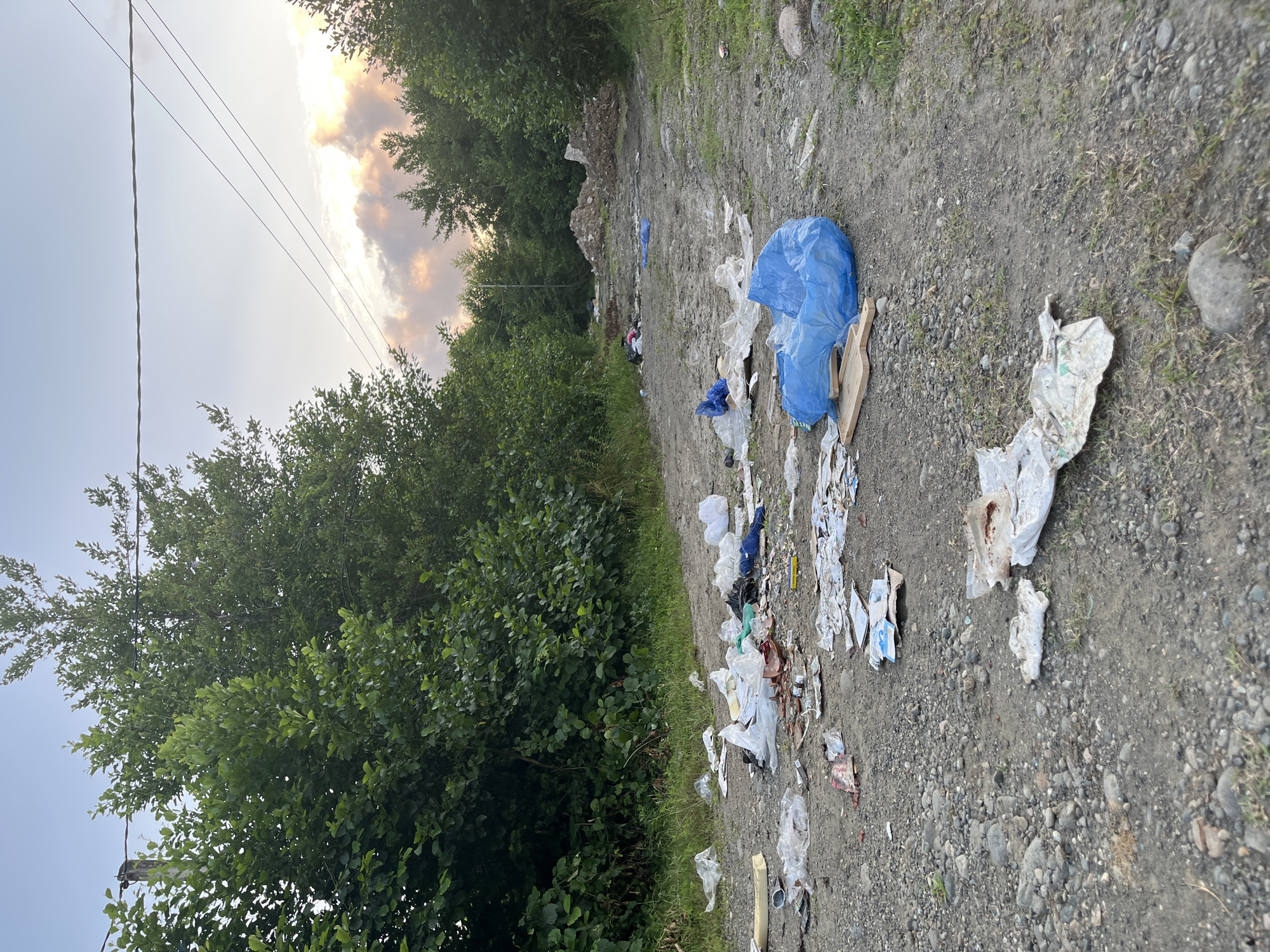 The image shows a dirt road covered in trash. There are plastic bags, food wrappers, and other debris scattered across the road and on the sides. Trees line the road, and there are power lines overhead. The sky is overcast with some clouds. The scene appears to be in a rural or undeveloped area.