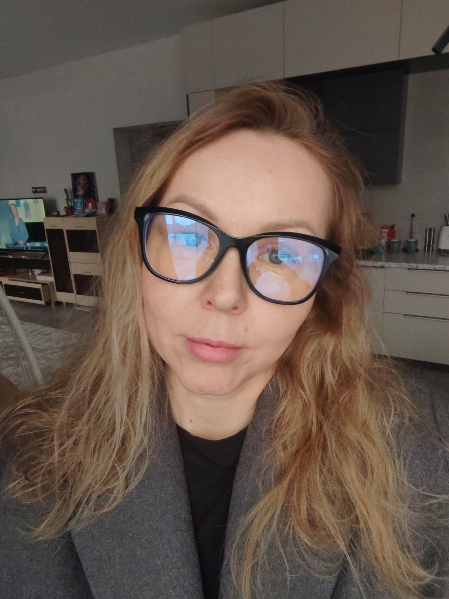 A woman with blonde, slightly dishevelled hair is wearing large, black-framed glasses. She is wearing a grey coat over a black top. The background shows a home interior with a television, shelving unit, kitchen cabinets, and various kitchen items. Based on the background elements, the location may be residential but there is insufficient information to verify the city of Chaikovsky, Russia.