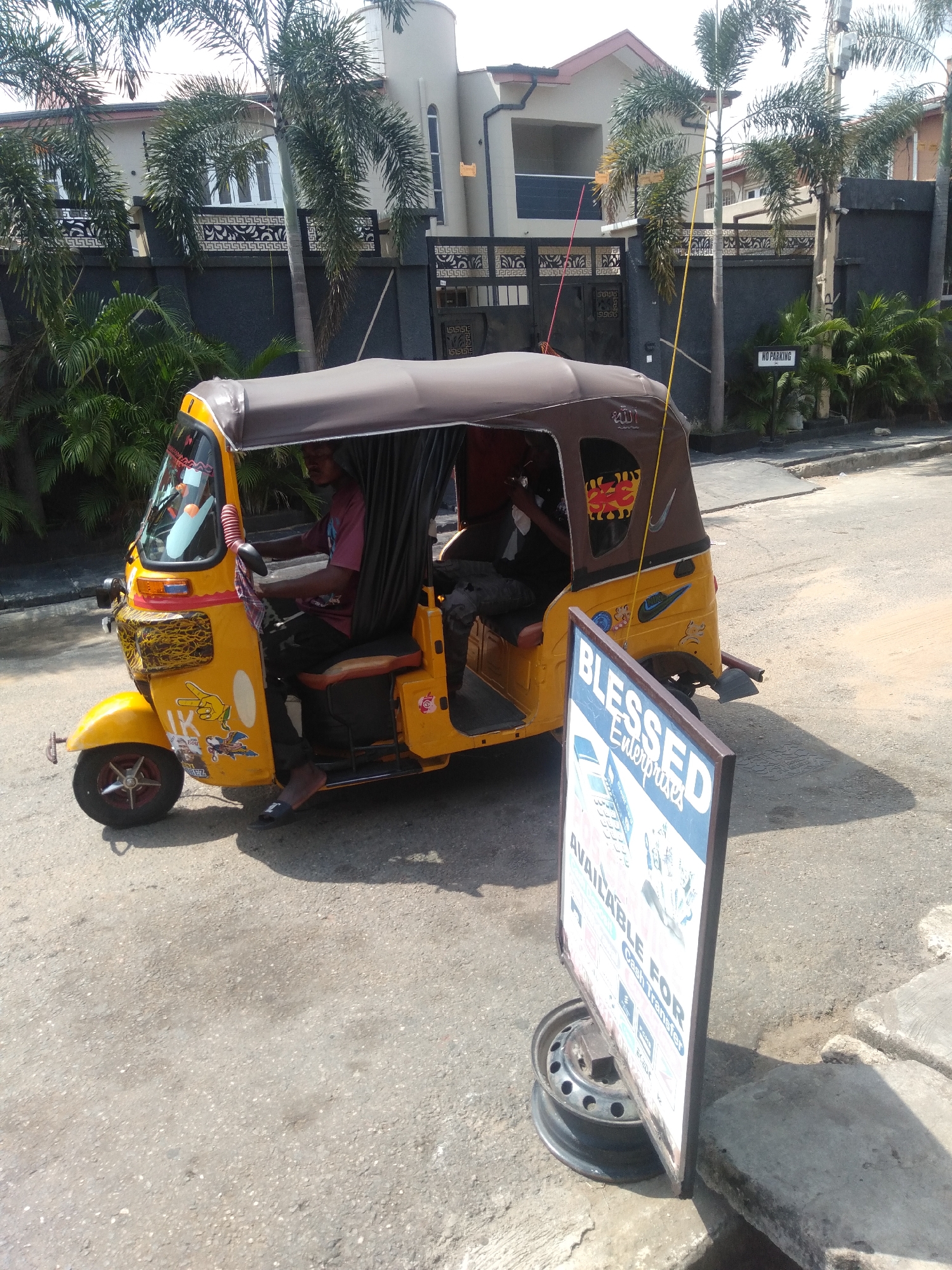 A yellow, three-wheeled motor vehicle, often referred to as a "keke" or "tuk-tuk," is parked on a paved road in Shangisha, Nigeria. The keke has a brown canopy and black curtains inside. A man is seated in the driver's seat. Behind the keke, a sign with the words "Blessed Enterprises" is visible, propped up by a car wheel rim. In the background are trees, a gray fence and a building with a white facade. A "No Parking" sign is fixed on the wall behind the vehicle. The environment suggests an urban setting on a sunny day.