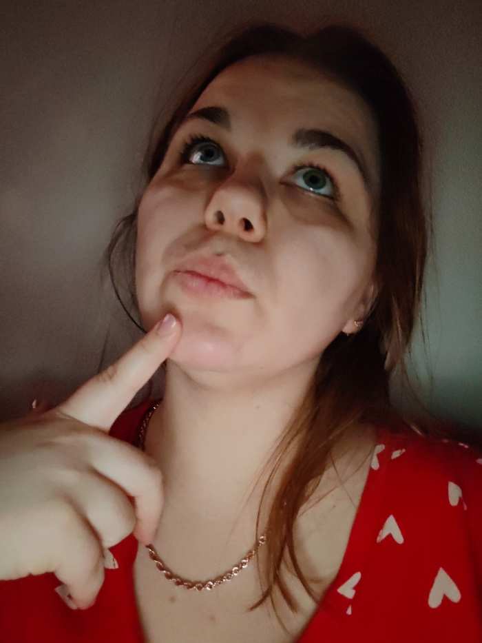 A woman in a red shirt with small white hearts is looking upwards with a thoughtful expression. Her index finger points to her chin. She wears a delicate gold chain necklace and small hoop earrings. She has light skin, green eyes, and brown hair pulled back loosely. The lighting is dim, creating shadows under her eyes and across her face.