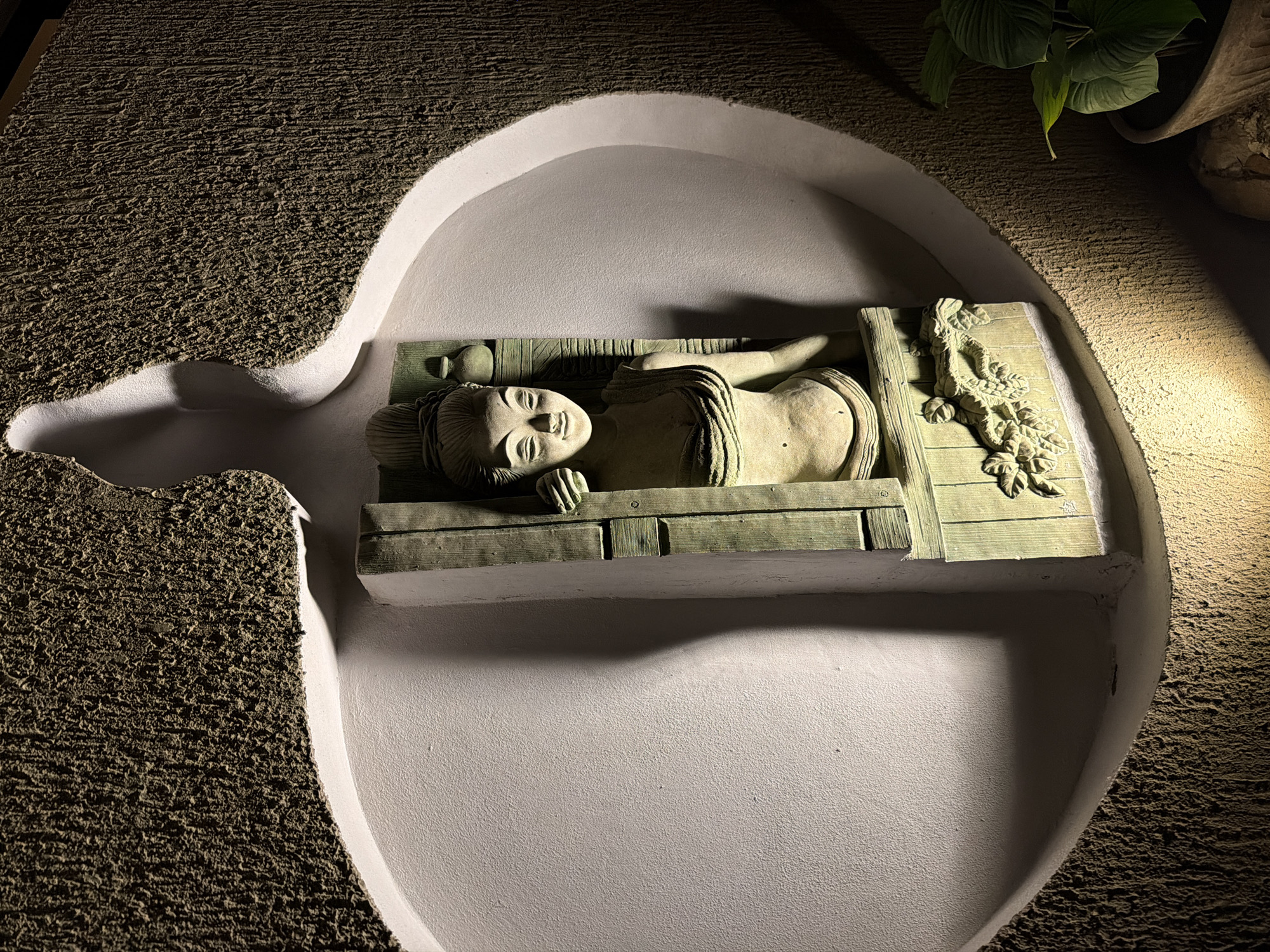 An illuminated sculpture depicting a woman in traditional Thai or Southeast Asian style is prominently displayed within a large, organic-shaped, smooth white niche on a dark, rough-textured wall in Ao Phai, Thailand. The sculpture, primarily in earthy tones with greenish patination, shows a woman with closed eyes and a slight smile, seemingly emerging from a carved frame. Her visible left hand rests on the frame, which features additional decorative carvings of foliage at its base. A small, decorative vessel is positioned on a ledge behind her head to the upper right. The scene is dramatically lit by spotlights from above and below, casting strong shadows within the niche and emphasizing the contrasting textures of the smooth inner surface and the granular outer wall. A potted plant with broad green leaves is partially visible in the lower right foreground.