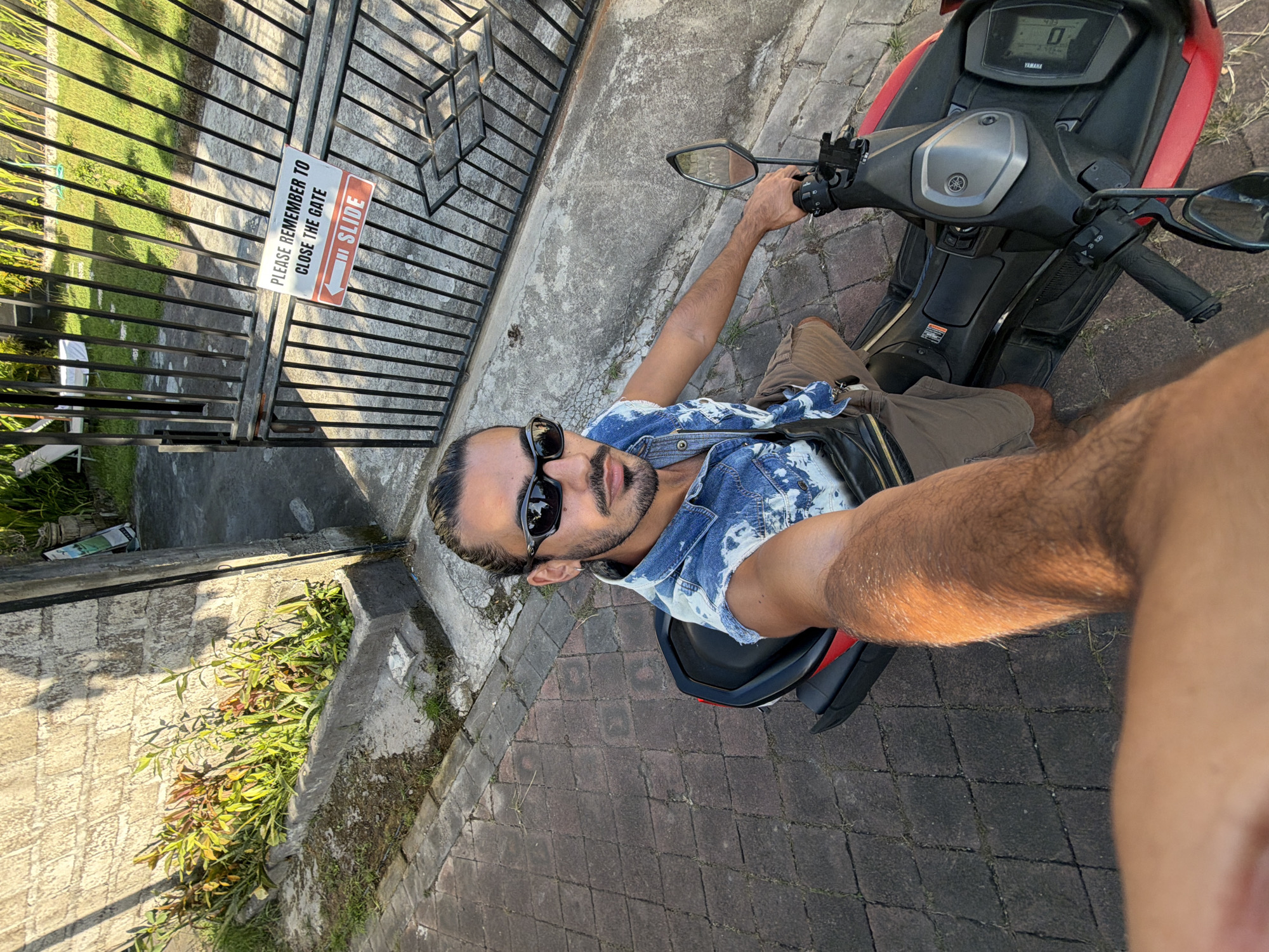 A man in sunglasses, a denim shirt, and tan shorts sits on a red and black Yamaha scooter, likely NMax model, on a brick-paved road in Kuta Selatan, Indonesia. The scooter's digital display reads "0". Next to an iron gate is a sign that says "PLEASE REMEMBER TO CLOSE THE GATE / SLIDE". The gate leads to a green area with plants. The scene appears to be outdoors during daylight.