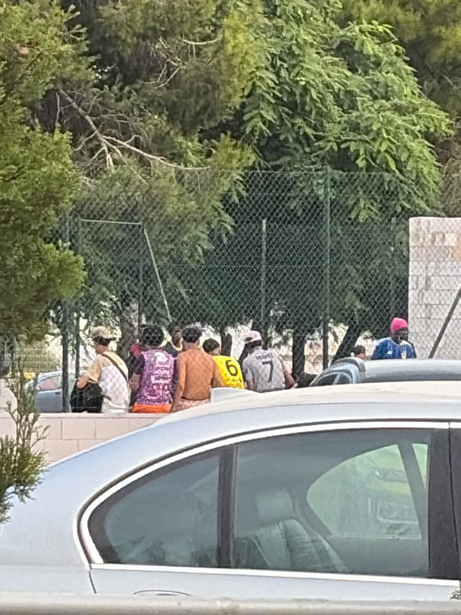 The image depicts a group of people gathered outdoors, some seated and some standing, behind a chain-link fence. The environment appears to be a park or recreational area, with trees and foliage visible in the background, and a concrete wall and structure on the right. A portion of a silver car is in the foreground, obscuring the lower part of the scene.
Specifically, the scene is located in Torrevieja, Spain, at the Municipal Sports Center's Padel courts. The people appear to be in casual attire. Some individuals are wearing t-shirts with numbers, including a "7" and a "6". One person is wearing a purple top with white patterns, and another is wearing a pink headscarf. The weather appears to be fair, with ample daylight. The overall impression is of a relaxed gathering, possibly after a sports event or during a break.