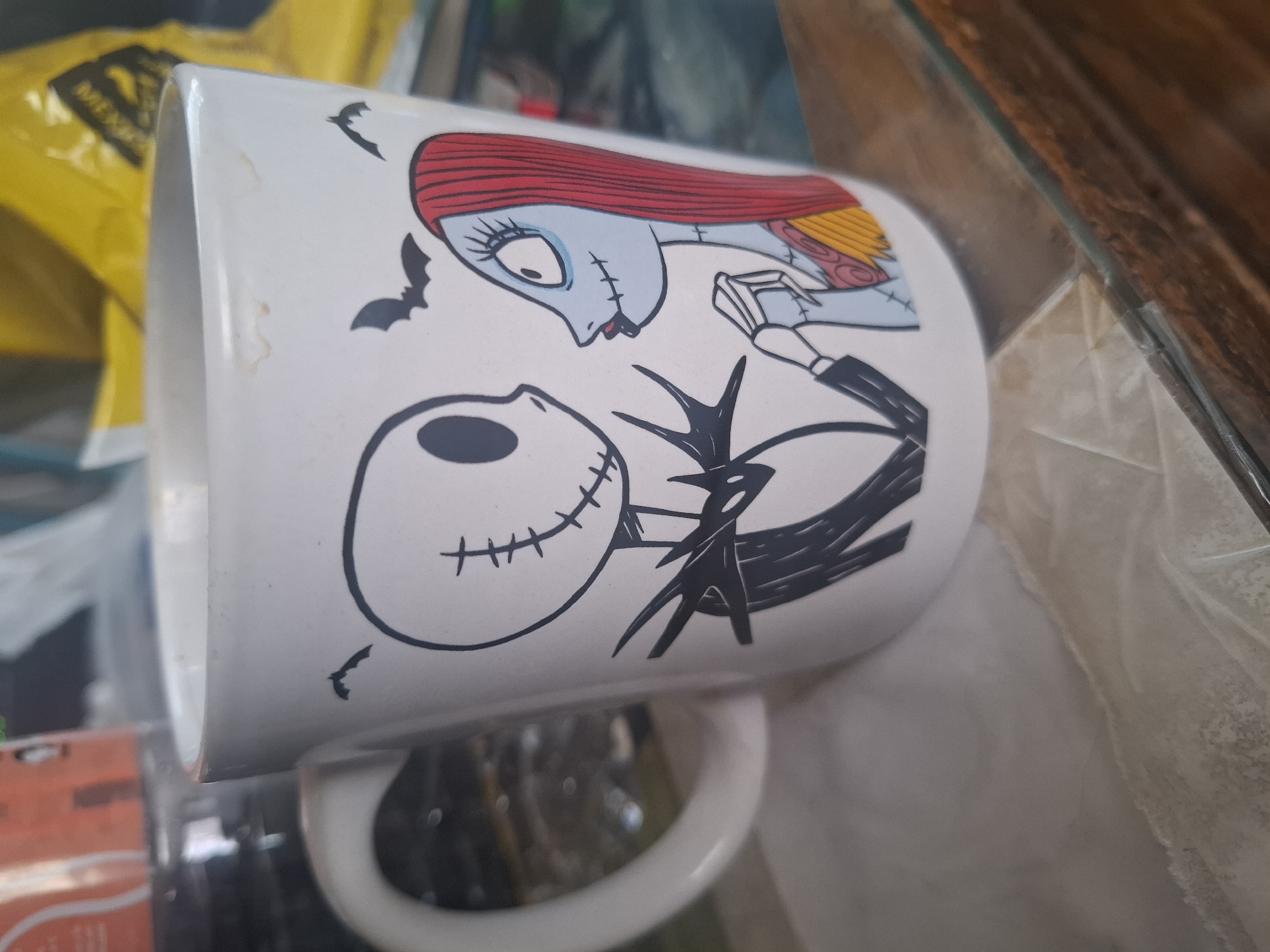 A white ceramic mug with a cartoon image of Jack Skellington and Sally from "The Nightmare Before Christmas" is sitting on a glass surface. The mug has a handle and is slightly stained. The background is cluttered with various objects, including a yellow bag. The location is Naucalpan de Juárez, Mexico, but there are no clues to place this image in the city.