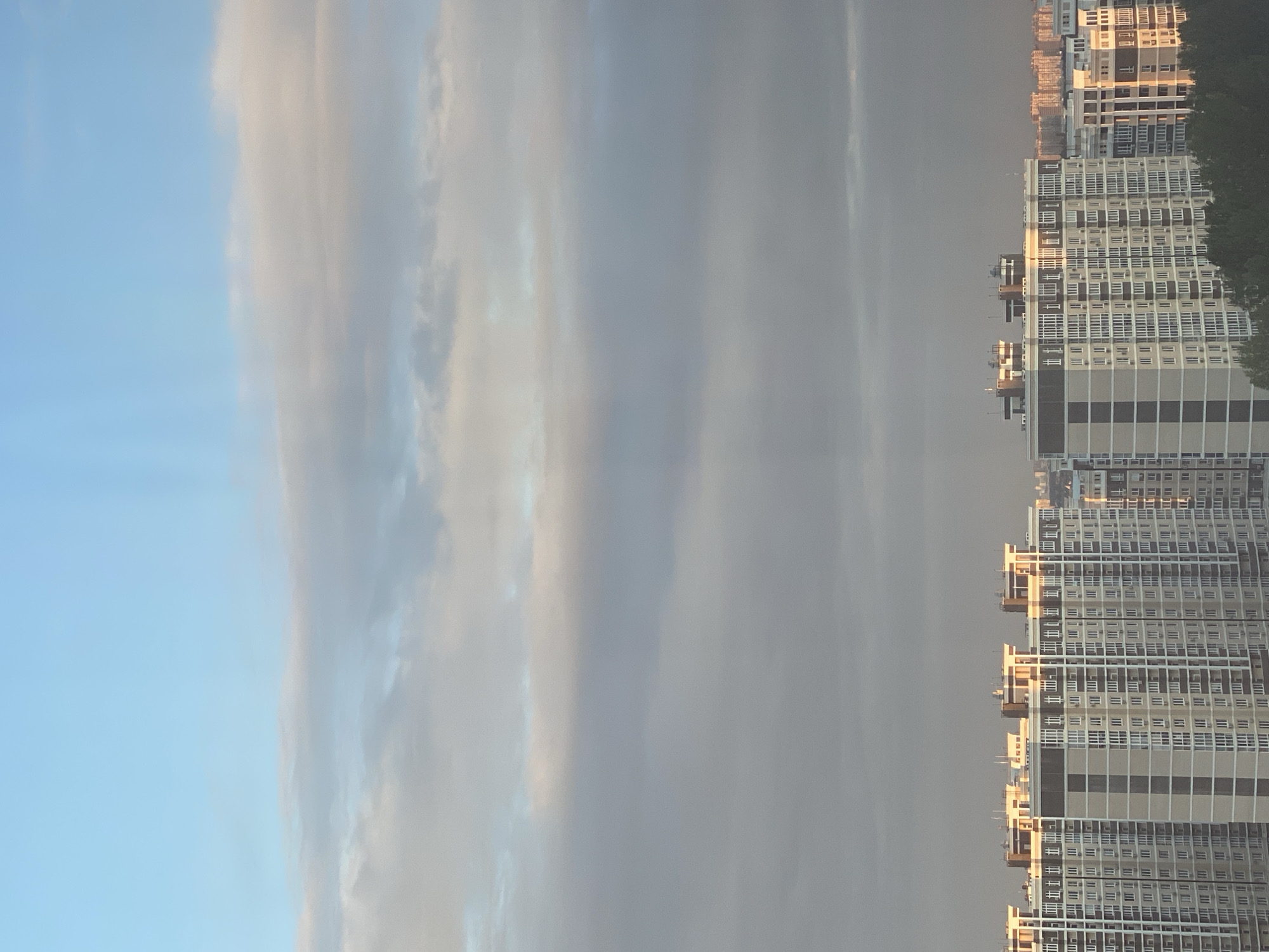 The image shows a city skyline at dusk. The sky is a gradient of light blue at the top, transitioning to grey and then dark grey as it nears the horizon. There are scattered clouds with soft edges, some illuminated by the setting sun, casting a warm glow on the upper edges. In the foreground, several tall, modern apartment buildings dominate the scene. These buildings are characterized by their numerous windows, visible balconies, and repetitive architectural patterns. The facades of the buildings exhibit a neutral color palette of beige, grey, and white. The setting sun casts long shadows and highlights the upper portions of the buildings with a warm, golden light, indicating the direction of the sun is from the right. In the far distance, more buildings are visible, appearing as a softer silhouette against the hazy sky. Dark green trees are present at the bottom edge of the image, partially obscuring the base of the apartment buildings. No people are visible in the frame. The scene depicts a typical urban residential area as daylight fades.