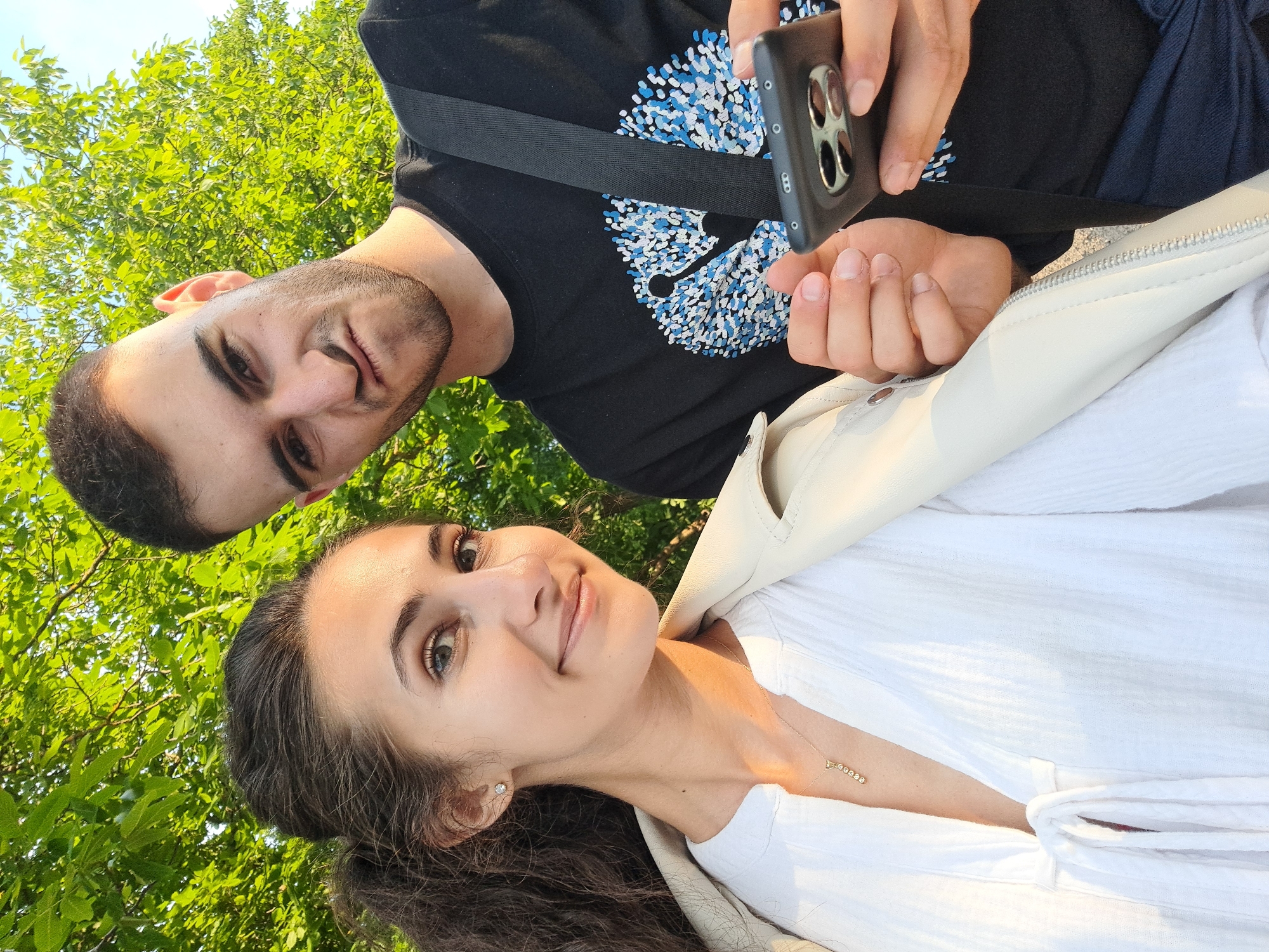 A medium shot selfie is taken outdoors with a young couple. The woman is on the left, facing the camera with a slight smile. She has dark, curly hair tied back in a ponytail and is wearing a white blouse with a tie at the neckline and a light-colored jacket. A delicate gold necklace with small diamonds is visible. The man is on the right, also facing the camera, with a playful, scrunched-up expression. He has short dark hair and is wearing a black t-shirt with a blue and white design and a black strap across his chest. He is holding a dark smartphone in his right hand, which is pointed towards the camera. The background consists of lush green trees with sunlight filtering through the leaves, suggesting a warm, sunny day. The scene appears to be in a park or a natural outdoor setting, possibly in the city of Lokorsko, Bulgaria, given the foliage and bright sky.