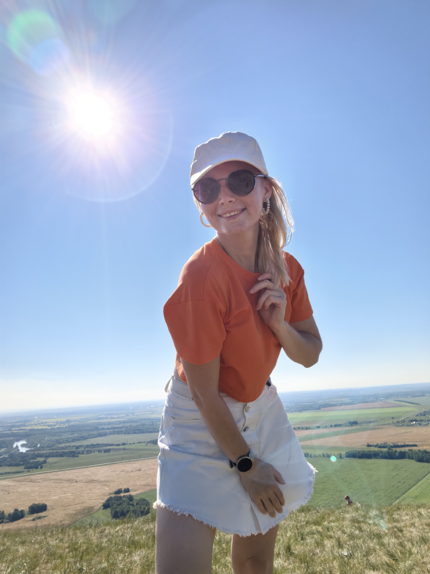 A woman stands on a grassy hill overlooking a rural landscape under a clear blue sky with the sun shining brightly. She is wearing an orange t-shirt, a white baseball cap, and a white denim skirt with a frayed hem. She also has on sunglasses and a smartwatch. The woman is smiling and touching her hair. The background shows fields, trees, a river, and a few distant buildings. The location is identified as the city of Shikhan, Russia.