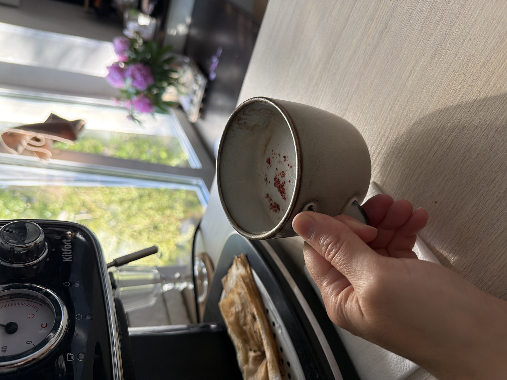 A hand holds a light-colored ceramic mug filled with a milky beverage, sprinkled with small red flakes. The mug is on a light wooden surface. To the left, a black espresso machine, labeled "Kitfort," is partially visible. A plate with folded, thin pastries sits on the espresso machine's drip tray. In the background, sunlight streams through a window, illuminating a bouquet of pink flowers in a vase and green foliage outside. The scene suggests a domestic setting, potentially a kitchen, in Chaikovsky, Russia.