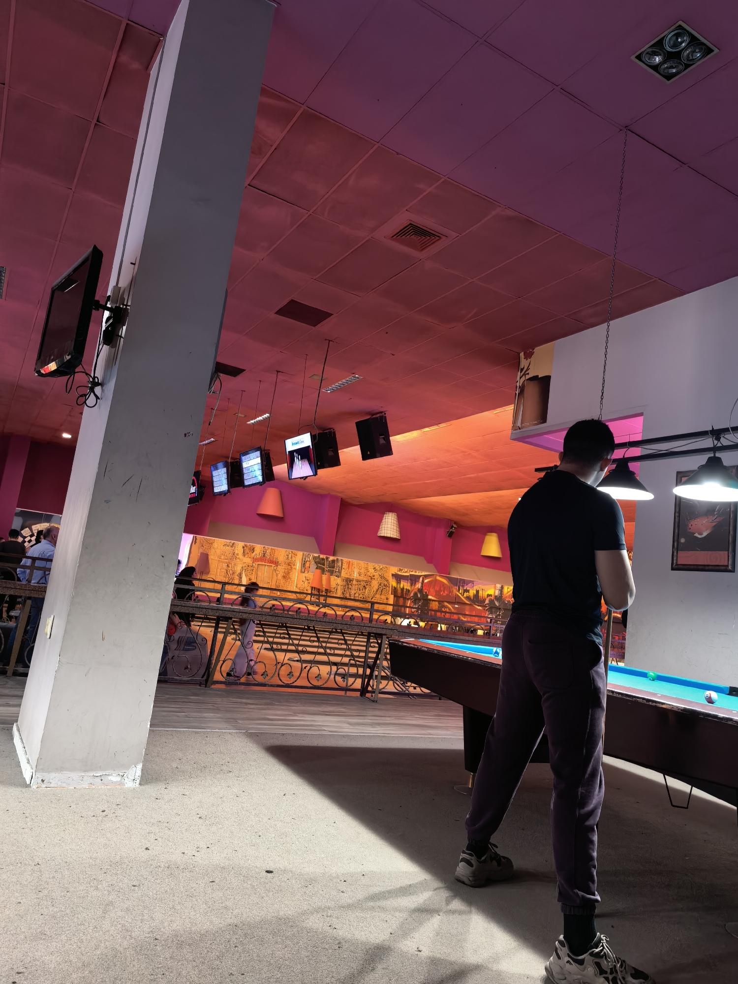 The image depicts an indoor scene in what appears to be a recreation center or club in Plovdiv, Bulgaria. A man stands at a pool table, ready to take a shot. The pool table is illuminated by hanging lamps. To the left, a large structural column supports mounted screens. A railing separates the pool table area from a lower level where other people are present, some seated. The color scheme is dominated by shades of pink and orange.