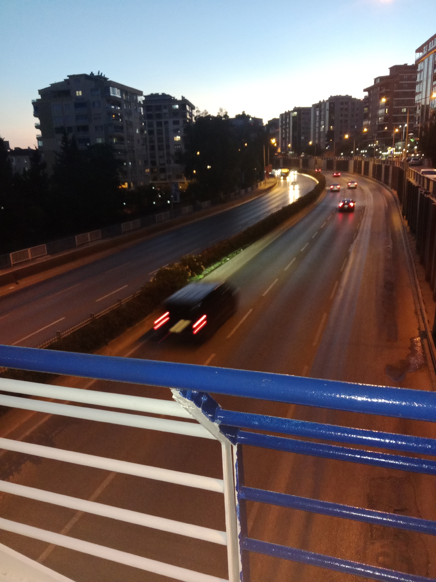 The image depicts a highway interchange at dusk in Karşıyaka, Turkey. The sky is a gradient of pale blue and light orange, indicating the transition from day to night. Multiple lanes of traffic are visible, with several cars in motion. Some vehicles' headlights and taillights are blurred due to motion, suggesting a long exposure photograph. The road surface appears wet, reflecting the streetlights and vehicle lights.
On either side of the highway, multi-story buildings are visible, typical of an urban environment. The buildings are dimly lit, with some windows showing interior lights. There are streetlights lining the road, illuminating the scene. Trees and foliage are also present, bordering the highway.
In the foreground, a blue and white railing is visible, suggesting the vantage point is from an overpass or a pedestrian bridge. The railing is made of horizontal bars and appears to be constructed of metal, painted blue and white.
The overall impression is of a busy urban transportation artery during the twilight hours.