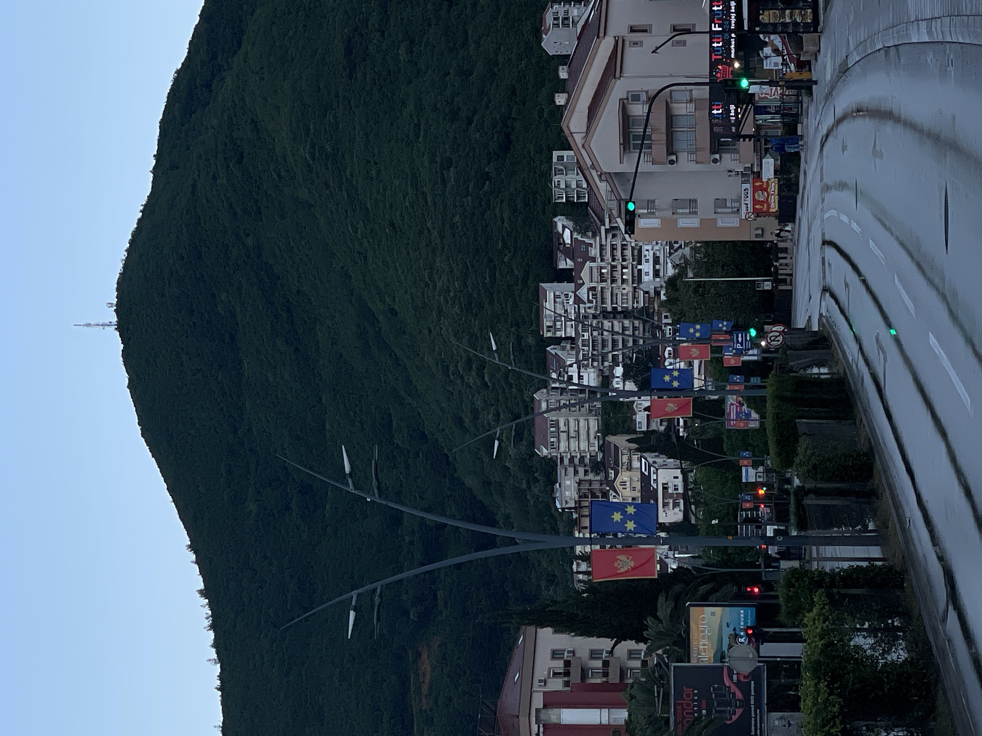 The image shows a view of a city street with a large, green mountain on the left side of the frame. The street is wet, suggesting it has recently rained. There are buildings of varying heights and designs on the right side of the street, with a mix of residential and commercial structures visible. Along the street are poles with flags, seemingly national flags of Montenegro and possibly European Union flags. Traffic lights are also visible, some showing green lights. Billboards and signs are placed along the street, containing commercial advertisements. The sky is light blue, indicating it could be early morning or late evening.