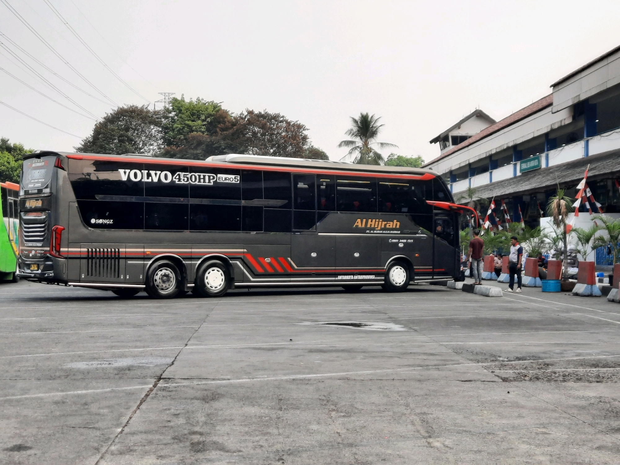In the city of East Jakarta, Indonesia, a dark gray tour bus with red and white accents is parked in a lot. The bus features the branding "VOLVO 450HP EURO5" and "Al Hijrah" in white lettering. The words "ANTARKOTA ANTARPROVINSI" are printed along the side of the bus, indicating intercity and interprovincial travel. Two men stand near the bus. One man, wearing a dark shirt and jeans, stands by the side of the bus. The other man, in a light-colored shirt and dark pants, stands further away, facing the bus. In the background, a building with a light-colored facade and a red-tiled roof is visible, decorated with Indonesian flags. The sky is overcast.