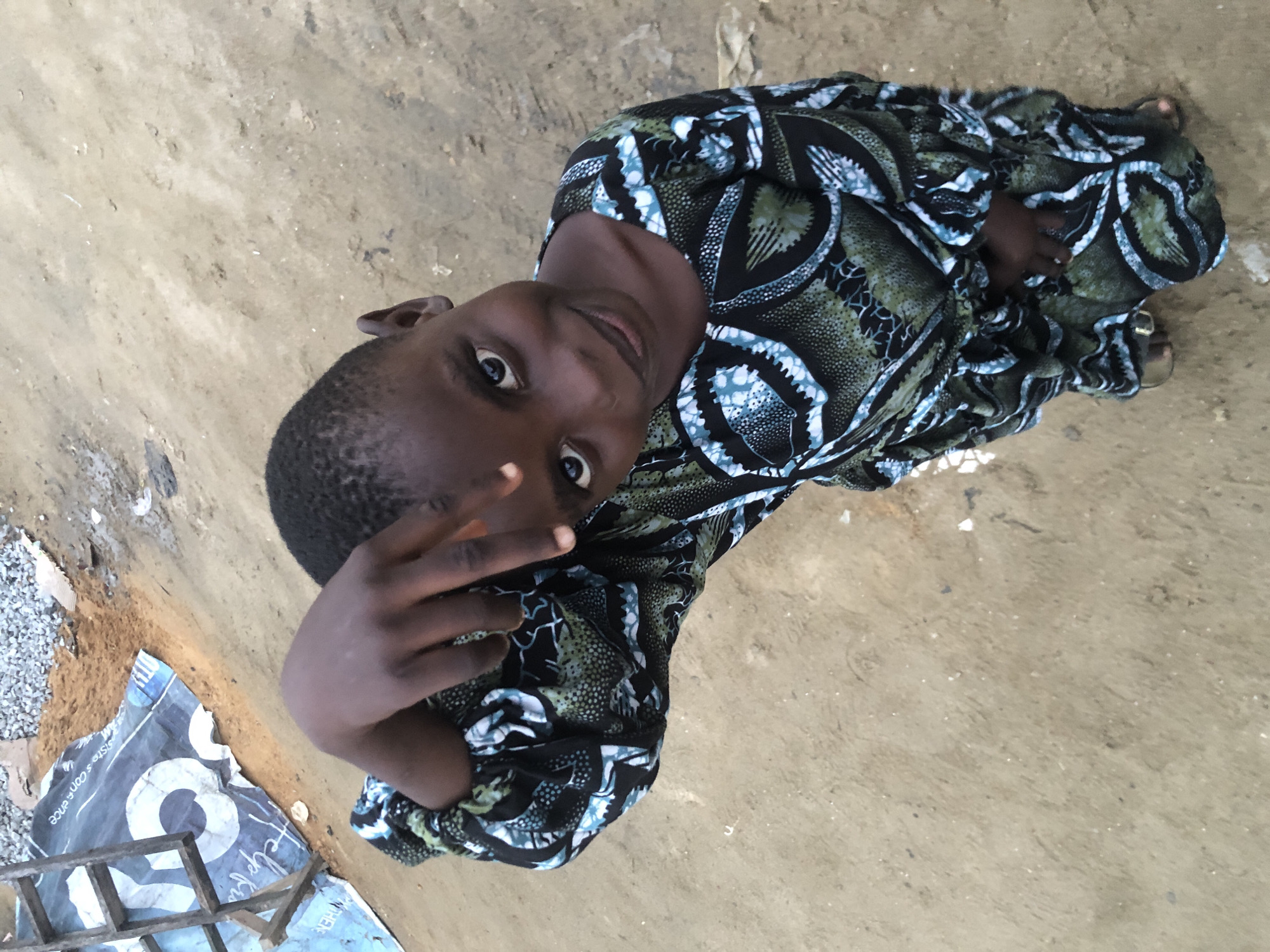 In Ado, Nigeria, a young girl stands on a sandy dirt surface. She is wearing a patterned dress in black, green, and blue. Her left arm is bent at the elbow, with her hand near her head, fingers pointing towards the camera. The girl is looking directly at the camera with a slight smile. In the background, a wooden structure, possibly part of a ladder, is visible next to a blue tarp with white circular markings. The ground appears to be a mixture of sand and gravel.