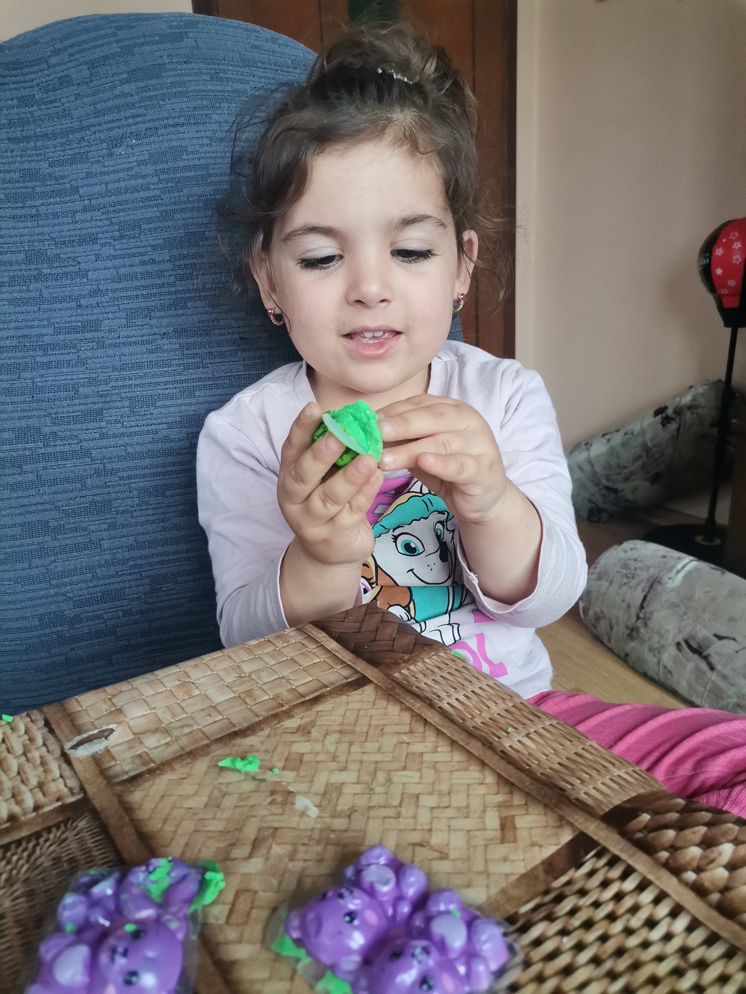 A young girl is sitting at a table, playing with green clay. She has brown hair pulled up in a bun and is wearing a long-sleeved shirt. The table has a wicker design and there are purple clay figures on it. In the background, there is a blue chair and a floor lamp. The location appears to be inside a home, the city and location being unknown.