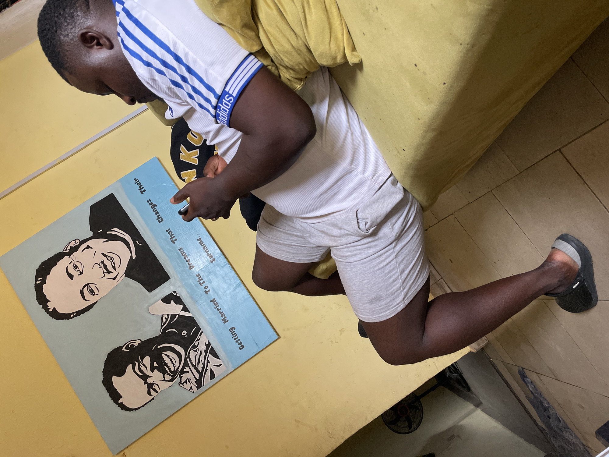 A man is lying on a yellow sofa, looking at a blue painting resting on a yellow surface. The painting depicts two black and white portraits of smiling men, with the text "Getting Married To The Dreams That Changes Their Surname..." The man is wearing a white t-shirt with blue stripes on the sleeves, and gray shorts. He is wearing a black slide on his right foot. The image shows a fan in the bottom right, and a tiled floor. The location is not explicitly stated as Jos, Nigeria, but the context suggests it could be a domestic interior in that region.