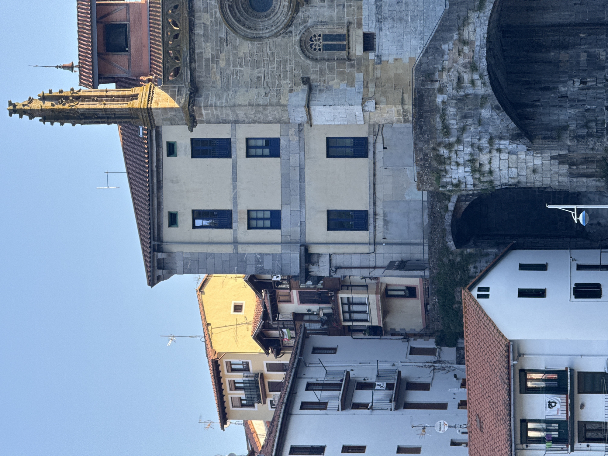 The image depicts a view of buildings in Ondarroa, Spain, likely near the river or coast, given the architectural style and presence of a bridge structure. The scene is dominated by a collection of multi-story buildings with red tiled roofs, varying in color and architectural details. On the right side of the image, a substantial stone structure, possibly part of a church or historic building, features a prominent gothic spire and a large circular window. Adjacent to it, a lighter-colored building with cream-colored walls and dark blue windows is visible. To the left, several white buildings with balconies and dark-framed windows are stacked in a typical residential configuration. One white building in the foreground has a red-tiled roof and several windows, including a distinctive pair of narrow windows at the top. The lower portion of the image shows the weathered stone arches of a bridge or elevated walkway, overgrown with some vegetation. The sky is clear and blue, suggesting daylight, and the lighting indicates it might be late afternoon or early morning, casting some shadows and highlighting the textures of the stone and building facades. No people are clearly visible in the frame.