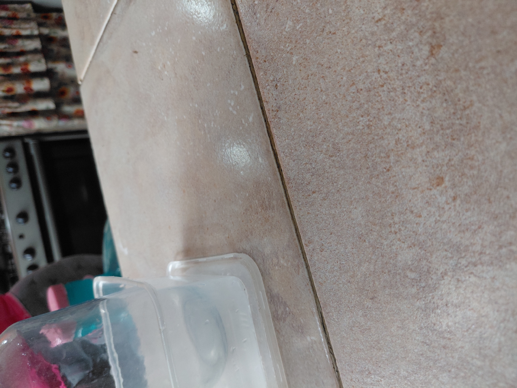 The image presents a close-up, horizontally oriented view of two distinct tiled surfaces and a partially visible translucent plastic container. On the left, a light beige tile with a glossy finish reflects light, showing faint white marks and possibly condensation or residue. This tile meets a darker, matte beige tile on the right, which exhibits a rough, granular texture with speckled patterns. A thin, dark grout line separates the two tiled sections.
In the lower-left foreground, a clear or translucent plastic container is visible, containing some liquid or moisture and displaying reflections on its surface. Its rim or lid is sharp-edged, suggesting a storage container. The background on the far left is heavily blurred, revealing stacked items with a floral or patterned design, possibly textiles or paper, and a dark object beneath them with circular controls, which may be part of a kitchen appliance like an oven or stove.
No specific activity is taking place, and there are no people or visible text. The scene depicts an indoor domestic environment, likely a kitchen or utility area, consistent with a home setting in a location such as Santa Ana, Venezuela. The time of day or external weather conditions cannot be determined from the image.