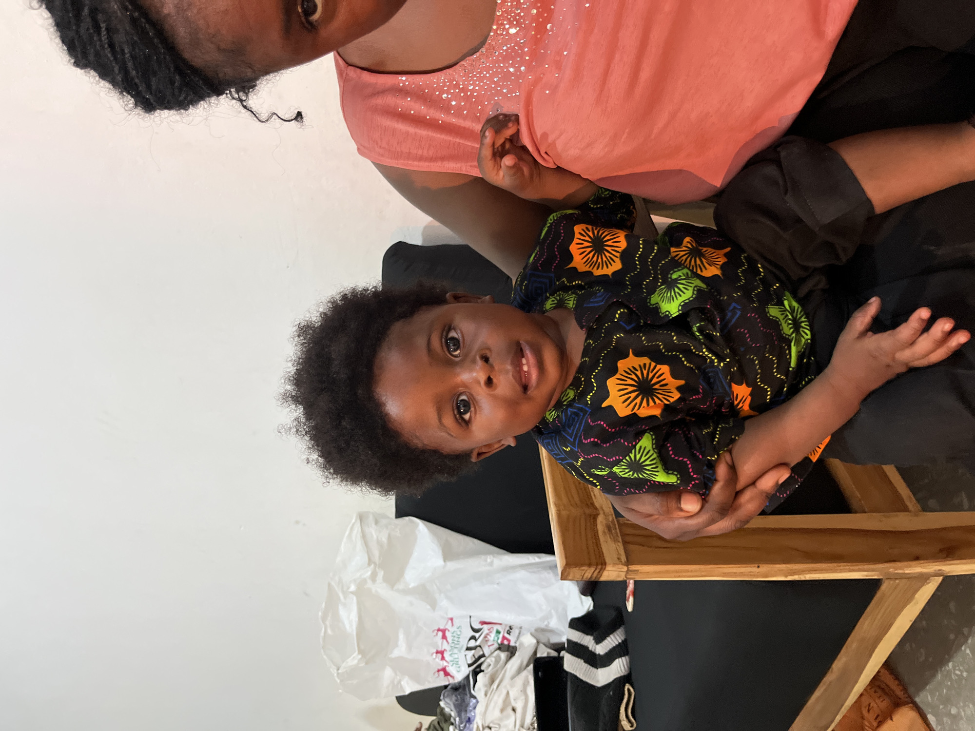 In Gura Top, Nigeria, a close-up, low-angle shot features a Black child lying across the lap of an adult. The child, with dark, curly hair and large, expressive eyes, looks directly at the camera. They are wearing a black t-shirt with a vibrant, abstract pattern of orange, green, and pink floral-like shapes. The adult, wearing a pink t-shirt adorned with scattered rhinestones, holds the child. The scene appears to be indoors, with a plain white wall in the background. To the left, a crumpled white plastic bag is visible, and some clothing items are piled on the floor. A wooden chair is partially visible in the foreground. The lighting suggests it is daytime.