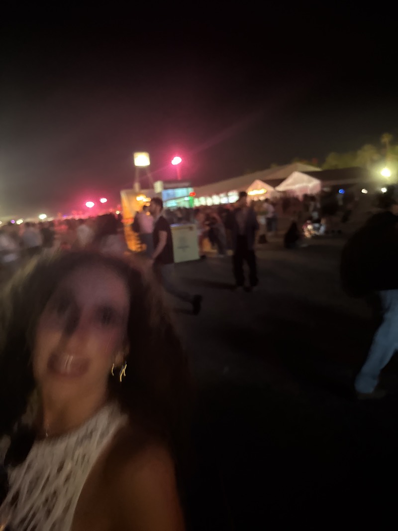 A blurry photograph taken at night in Indio, United States, depicts a crowd of people. In the foreground, a woman is holding the camera, partially obscuring her face. She is wearing a white top and gold earrings. Behind her, a crowd of people is gathered in a public space. There are tents or structures visible in the background, illuminated by lights. Some people are walking, and others appear to be standing or sitting. The scene suggests an outdoor event or festival.