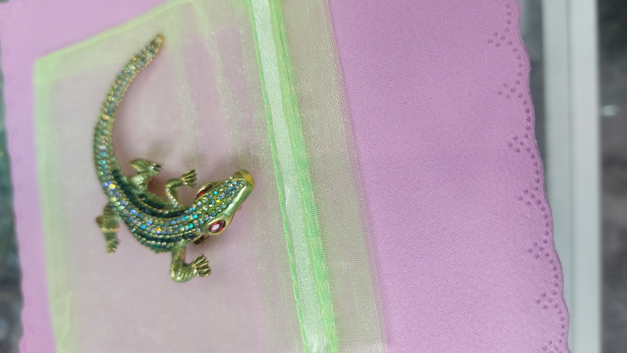 A close-up shot depicts a brooch in the shape of a reptile, possibly a lizard or alligator, adorned with green, clear, and red crystals. It rests on a layered surface: a light green sheer fabric is placed over a pale pink textured cardstock. A light green ribbon runs vertically down the middle of the frame. The scalloped edge of the pink cardstock is visible on the right. The setting appears to be indoors, and a small portion of a white frame or surface is on the far right. There is no indication of the location being Novosibirsk, Russia.