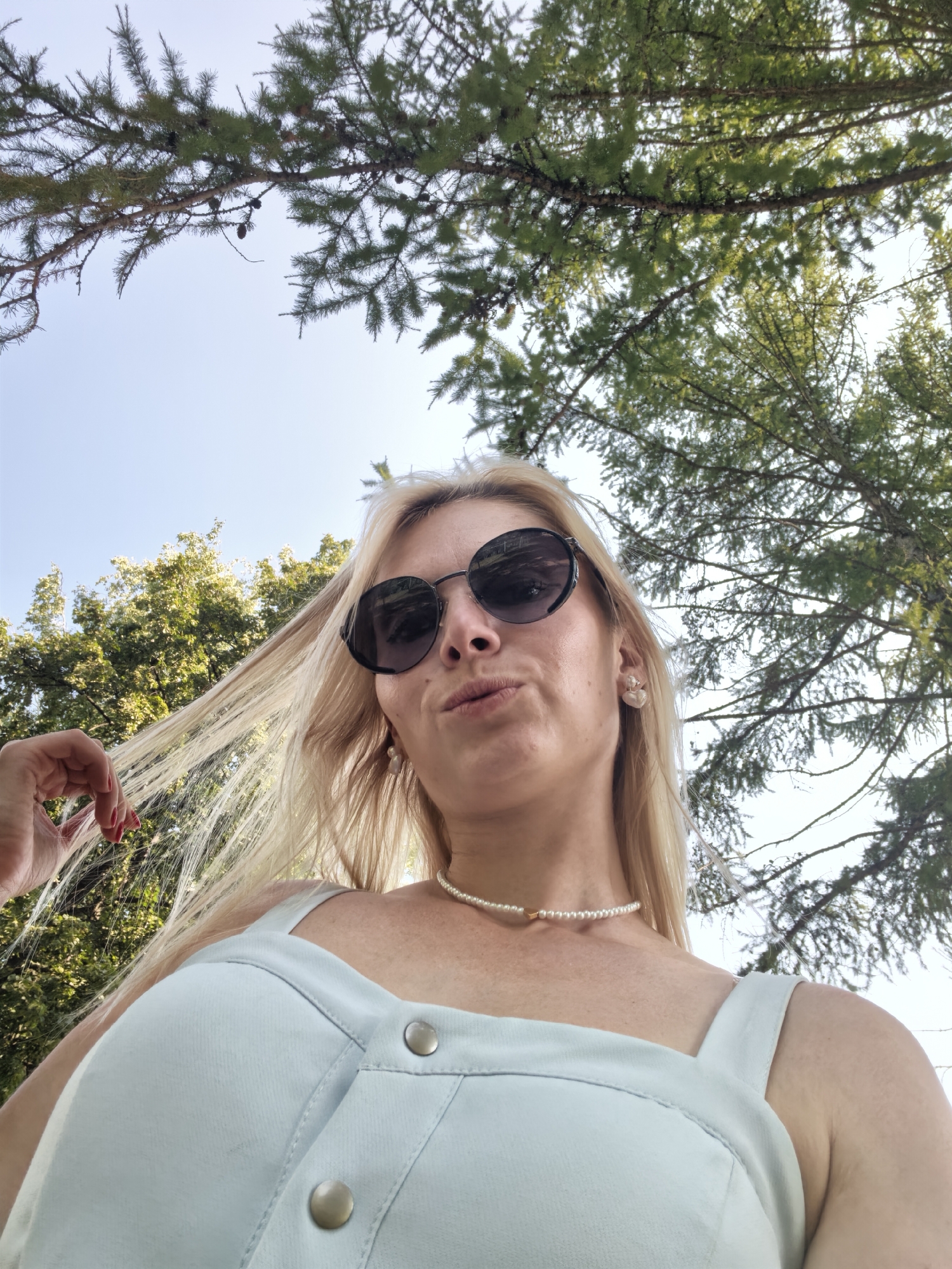 An outdoor, low-angle, waist-up, medium shot shows a fair-skinned woman from the chest up, looking directly into the camera. The woman has long, blonde hair, which is being played with by her right hand. She is wearing large, black, round sunglasses and a light blue top. Her hair is illuminated by the sun. She has on a delicate pearl necklace. The background is made up of a clear blue sky and the branches of green coniferous trees. The image appears to have been taken in Sterlitamak, Russia, on a sunny day.