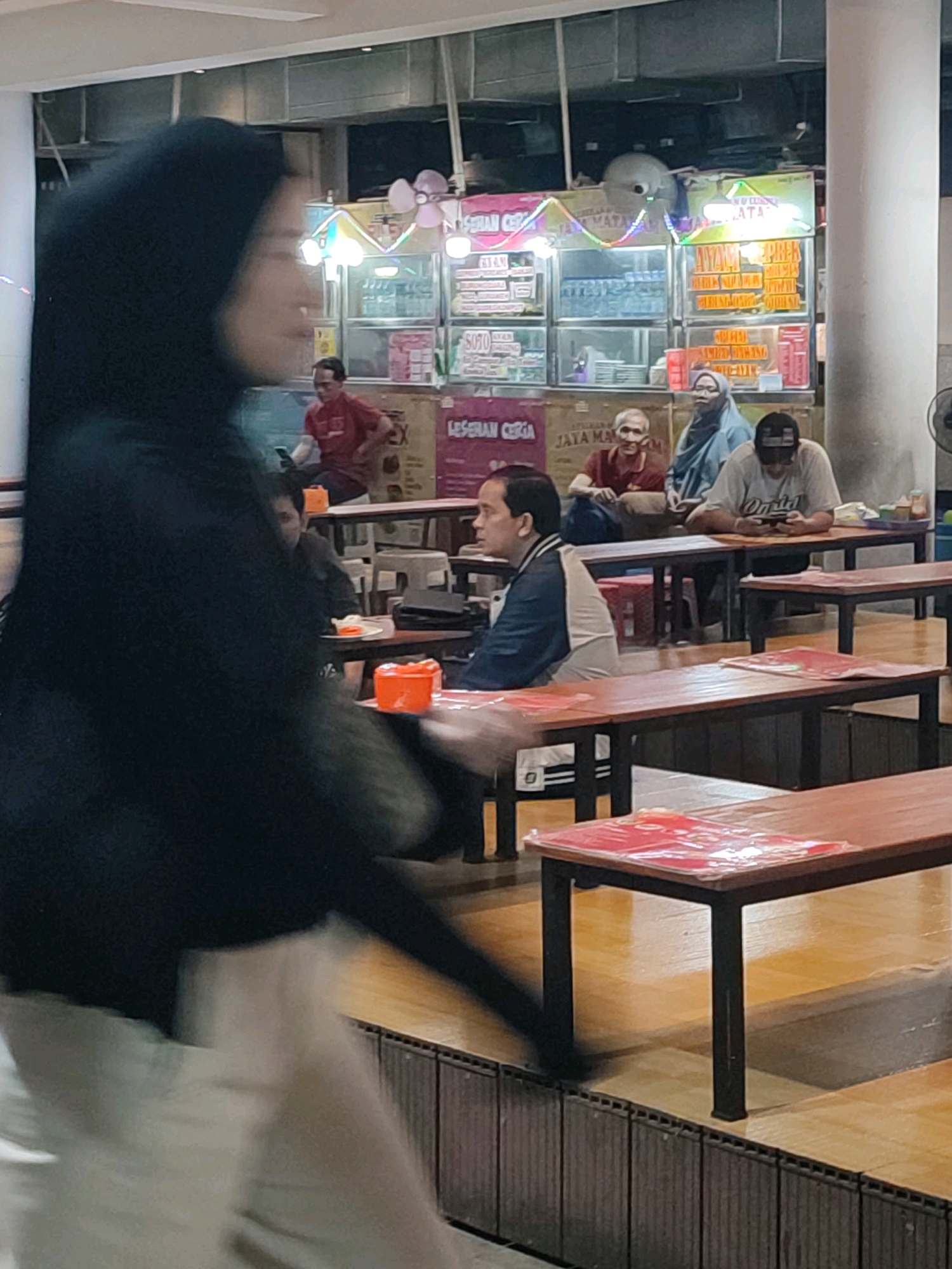 The image depicts an indoor food court or restaurant area, likely located in Yogyakarta, Indonesia, given the signage in Indonesian. In the foreground, a person wearing a black hijab and white clothing is out of focus and moving, obscuring a portion of the scene.
The background features several dining tables with wooden tops and black metal legs. Some tables are covered with red placemats or menus. There are several people seated at the tables.
On the right side of the image, a man wearing a gray t-shirt and a black baseball cap is looking down at his phone while seated at a table. To his left, a person wearing a blue hijab is also seated. Further back, a man in a red shirt is seated and looking at his phone.
Behind the diners, there are food stalls or kiosks with illuminated signage. One sign is partially visible with text that appears to be in Indonesian. The overall environment suggests a casual dining setting. The flooring is wooden, and the walls have a textured finish.