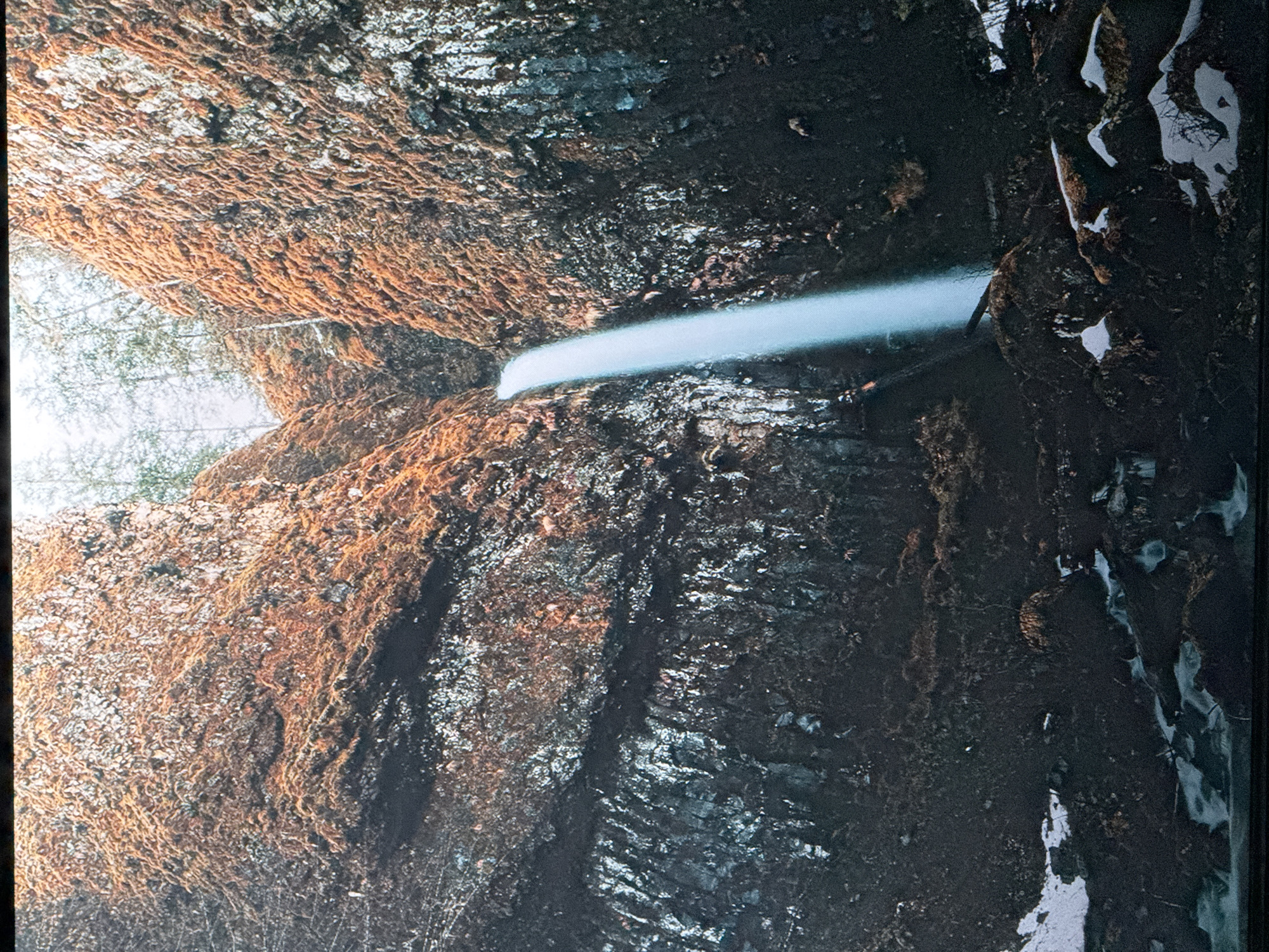 The image depicts a waterfall cascading down a moss-covered, rocky cliff face. The rocks are dark and textured, with patches of reddish-brown moss and lighter, wet-looking streaks. The waterfall itself is a bright, ethereal stream of white water, appearing to fall from a considerable height. At the top of the cliff, sparse trees with bare branches are visible against a pale, overcast sky. The base of the waterfall shows the rocky terrain and some patches of snow, suggesting a cool or wintery environment. There are no people or animals visible in the scene. The overall atmosphere is one of natural beauty and rugged wilderness. This scene is not located in the city of Saratov, Russia.