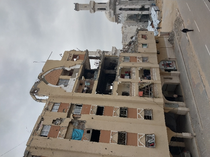An outdoor scene in Khan Yunis, Palestinian Territory, depicts extensive urban destruction under an overcast sky. In the foreground and mid-ground, a multi-story building, with upper floors showing features consistent with residential use and a ground-floor commercial unit identified by signage, exhibits severe structural damage. Its facade is heavily compromised, with large sections missing, exposed interiors, and numerous broken or absent windows. Evidence of fire damage, including blackening, is visible on interior walls and structural components on the middle floors. Some balconies still contain items resembling laundry. The ground floor features arched entrances and a commercial storefront displaying a red and white logo, "KFC," and "MAXIM" text. To the right and further back, a large mosque with a central dome and a tall, slender minaret also shows extensive damage, with portions of its structure appearing fractured or collapsed. Piles of debris, including corrugated metal, are visible near the mosque. The paved road in the foreground features visible lane markings and is bordered by scattered rubble and dirt. Four individuals are present: one person walks on the street in the foreground, facing away; another stands beneath an arched entrance of the damaged building, also facing away; and two smaller figures are visible in the mid-ground near the damaged mosque.