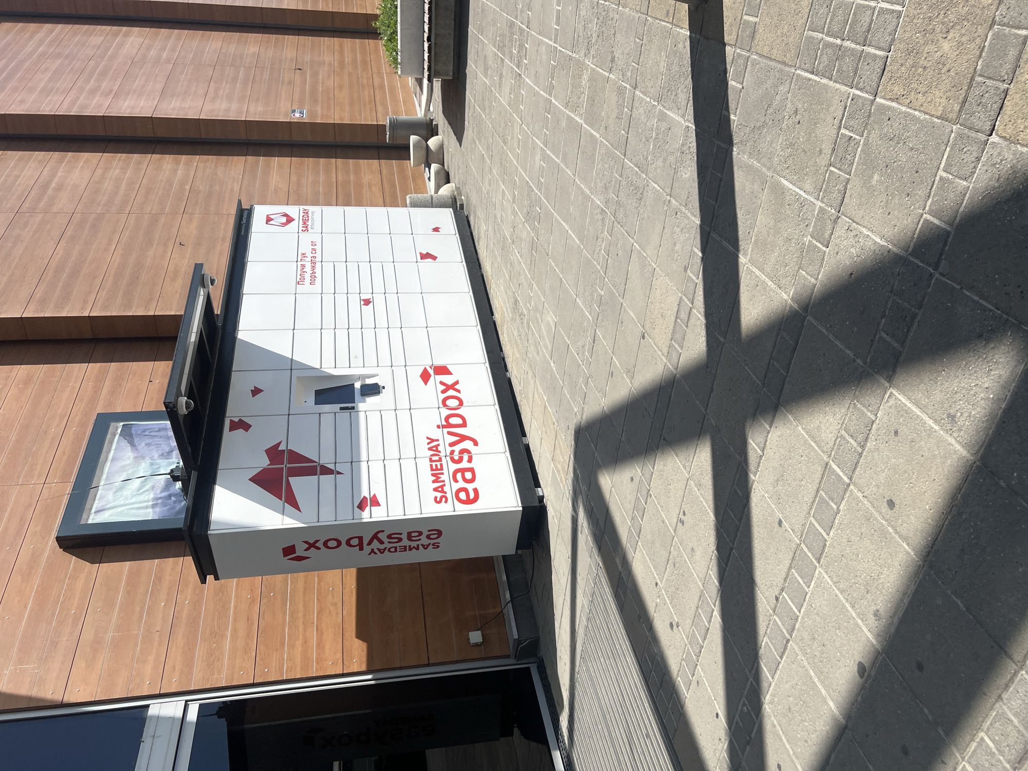 The image depicts an Easybox locker by Sameday, located in Burgas, Bulgaria. The locker is situated adjacent to a building with horizontal wooden-style paneling. A window is open on one side of the locker. The ground surface is paved with rectangular tiles. The lighting suggests daytime, with shadows cast on the pavement.