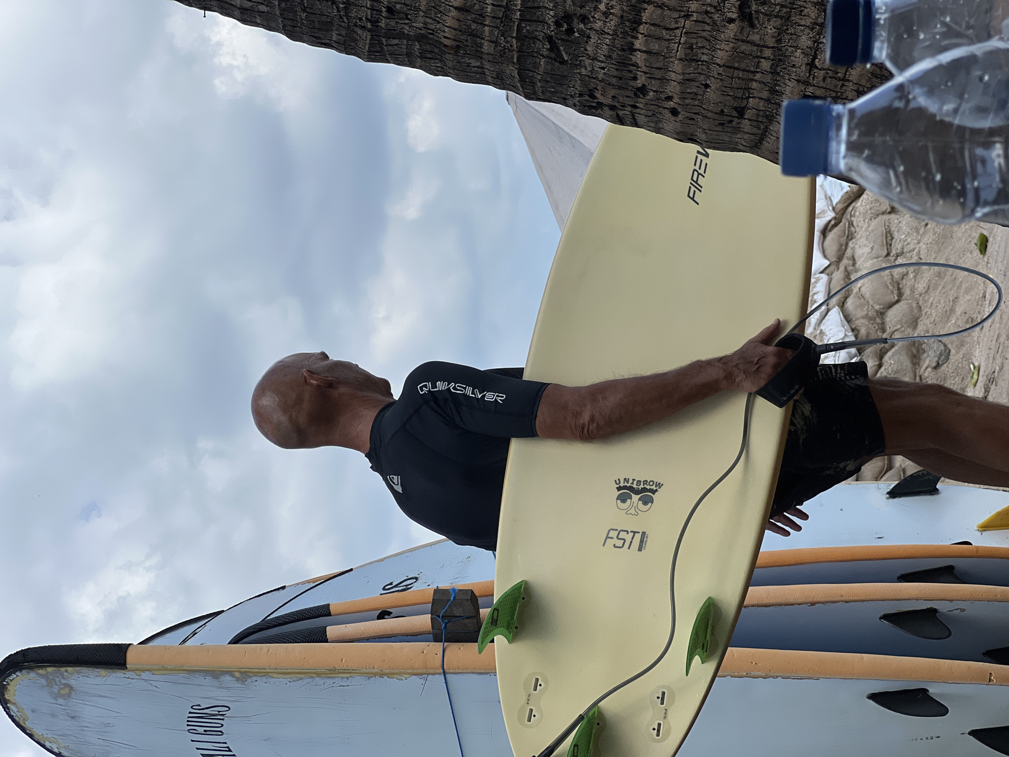 In Kuta, Indonesia, a bald, middle-aged man wearing a black Quiksilver rash guard and black shorts is standing with his back to the camera, holding a light yellow surfboard. He is looking to his right, towards a palm tree and two plastic water bottles in the foreground. The surfboard has green fins and a leash attached. Behind the man are several other surfboards, one of which has "CALI GUNS" written on it. The scene appears to be a beach, with sandbags visible in the background. The sky is cloudy.